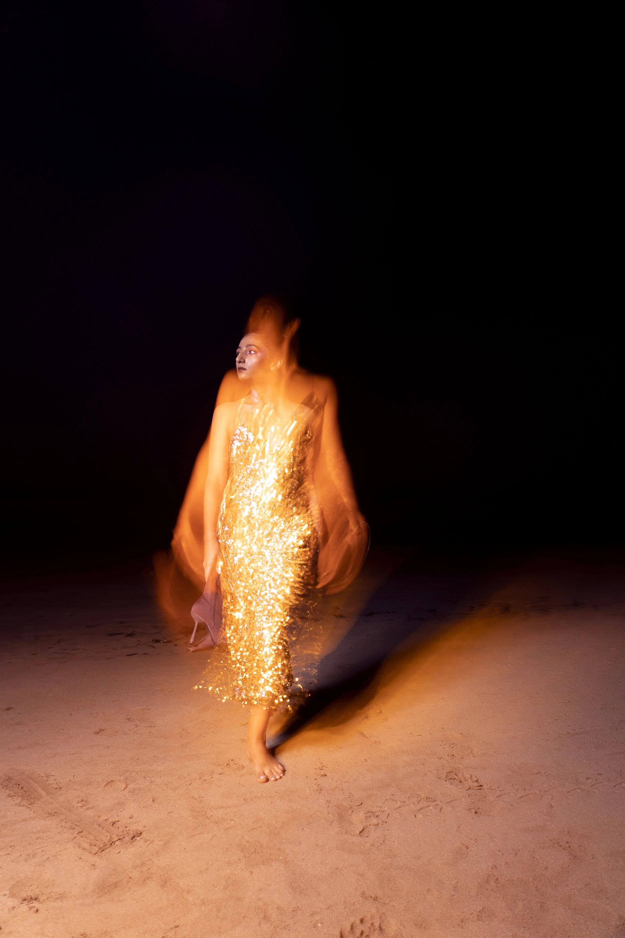 Woman in a sparkly gold dress standing or walking on a sandy surface at night, with motion blur and a dark background.
