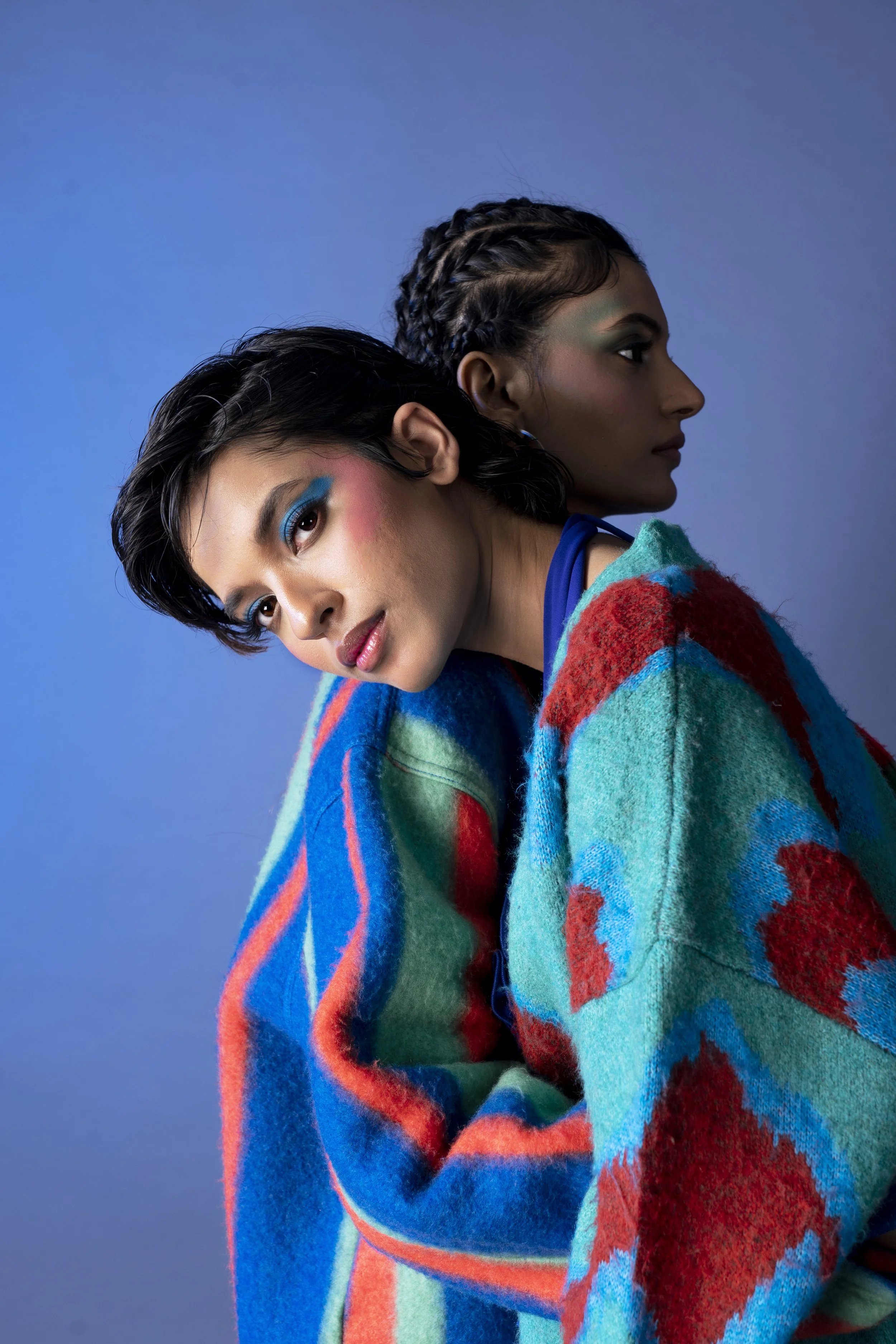 Two women with unique hairstyles and colorful makeup wearing a multicolored knitted sweater, posed against a blue background.