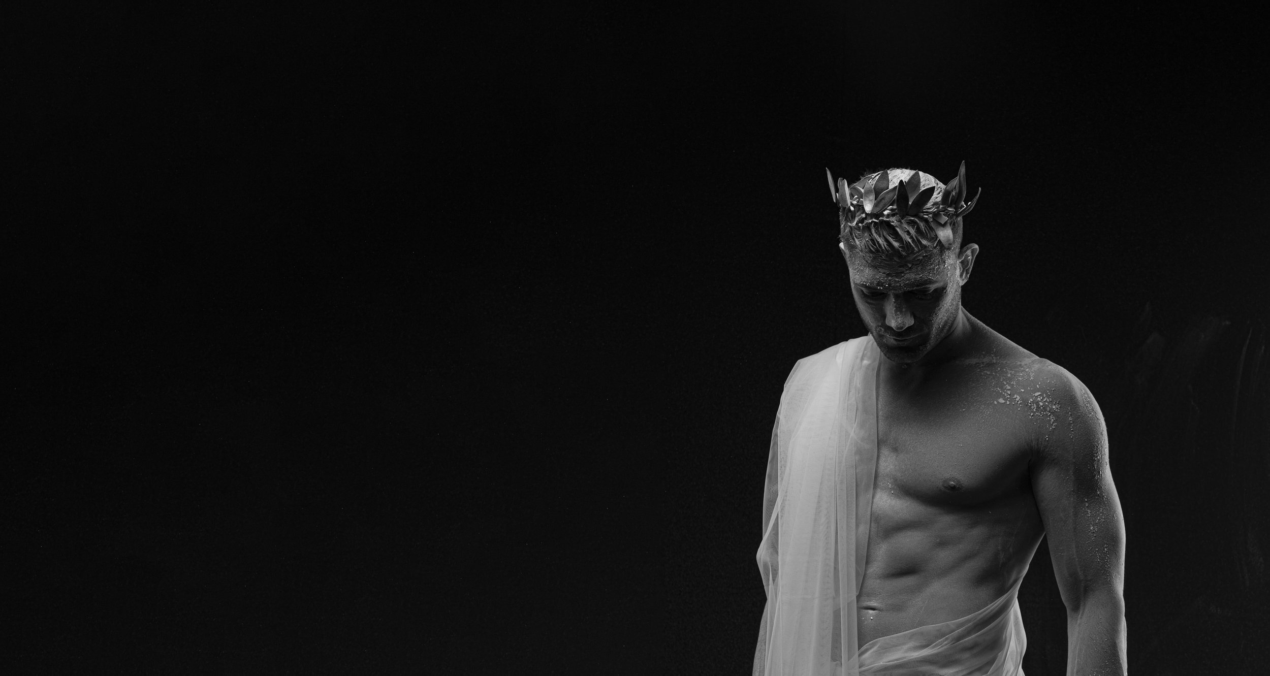 Black and white portrait of a young man with a laurel wreath on his head, wearing a toga draped over one shoulder, with a serious expression, against a dark background.