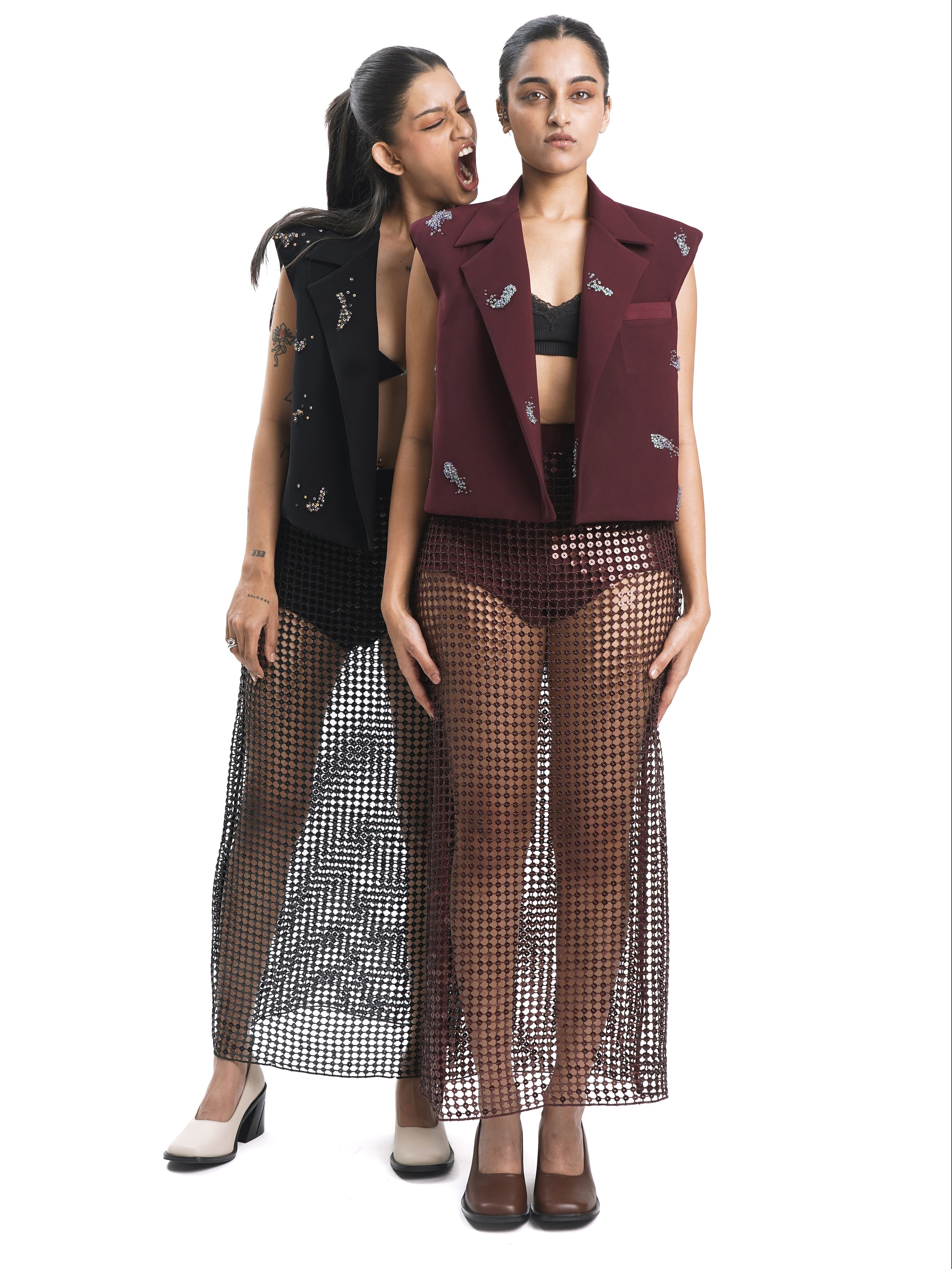 Two young women wearing stylish, edgy clothing made of mesh, with one woman whispering or yelling into the other's ear, who is standing with a neutral expression.