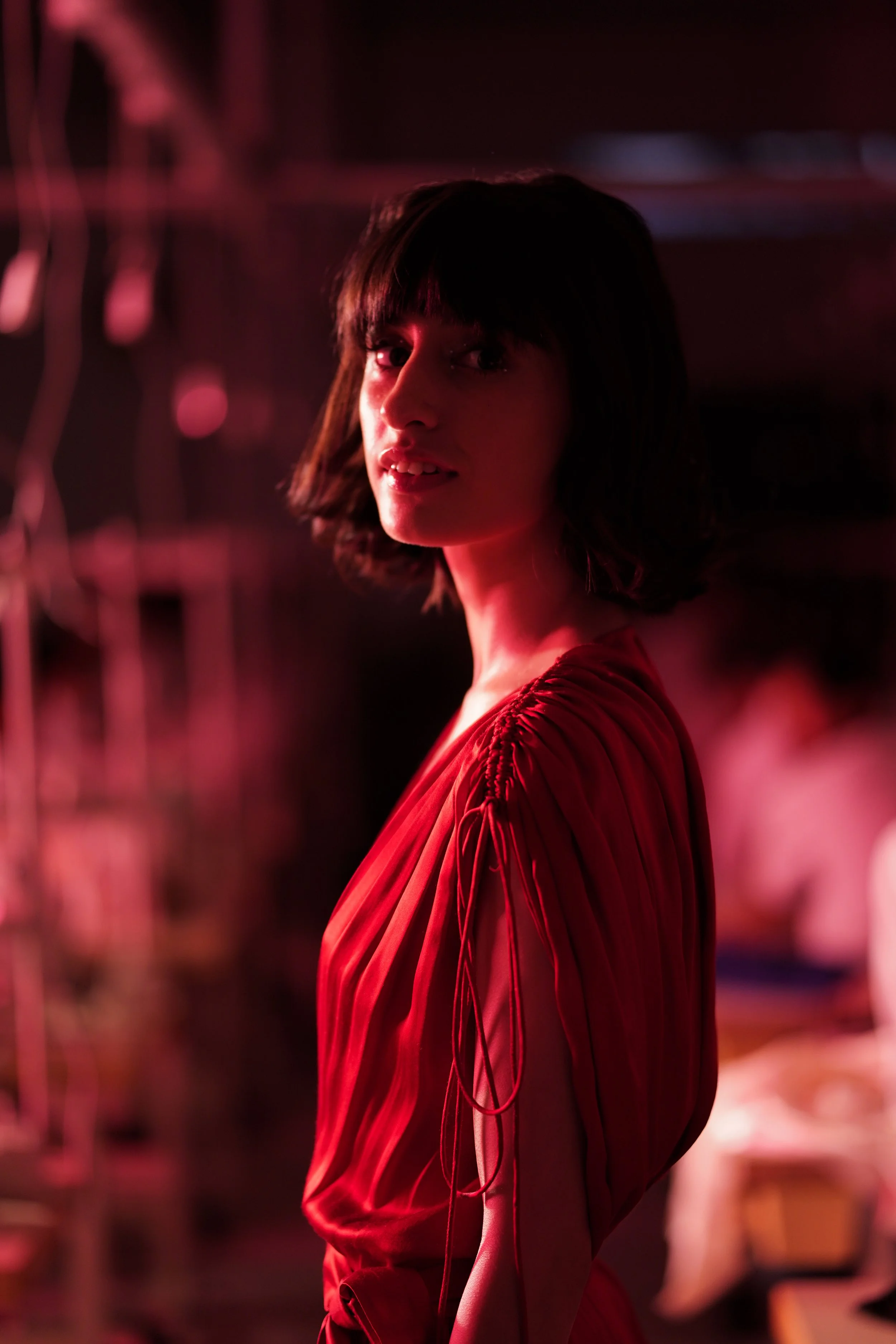 A woman with short dark hair wearing a red dress with gathered sleeves, illuminated by pinkish-red lighting in a dark environment.