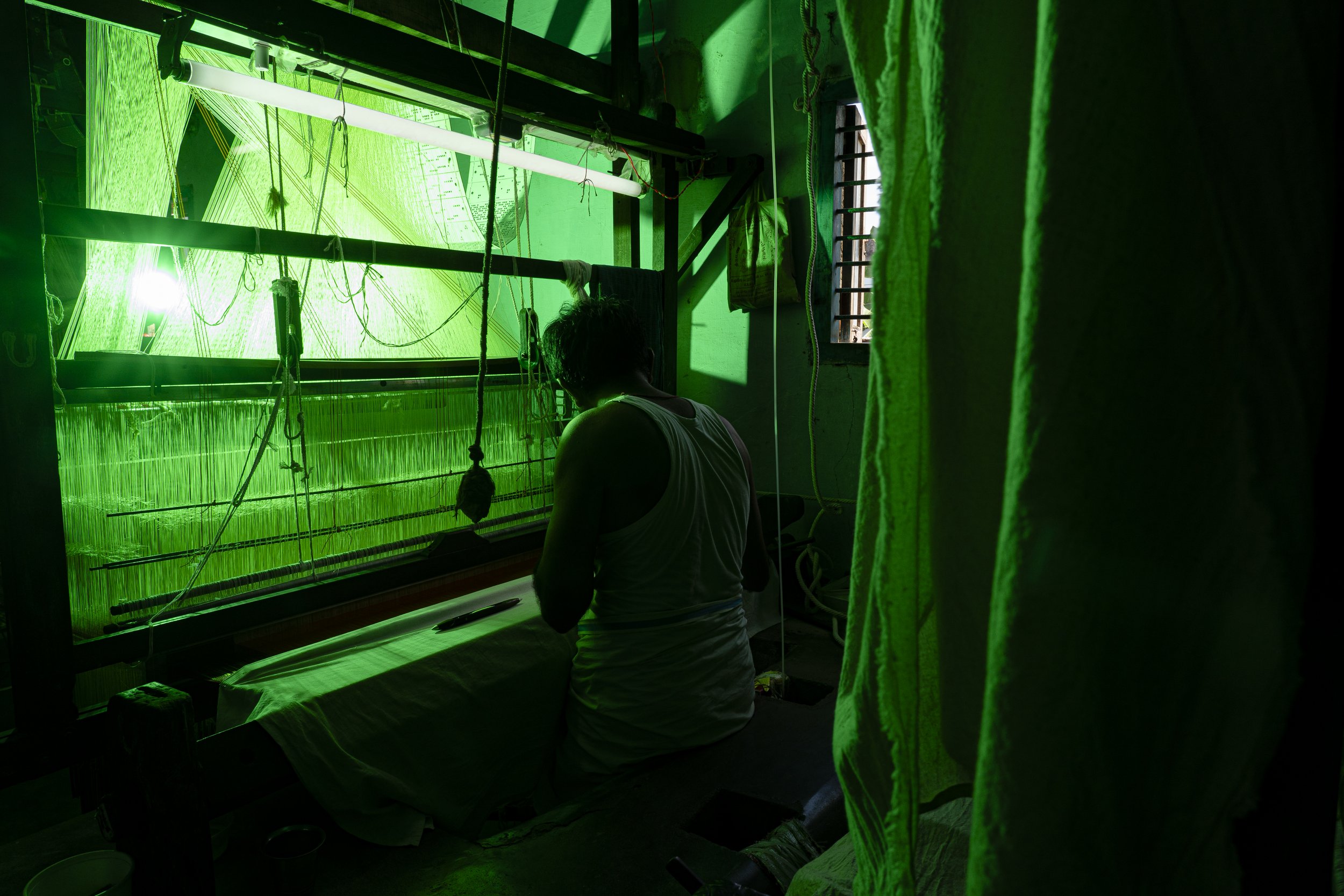 A person working on a loom in a dimly lit room illuminated by green light.