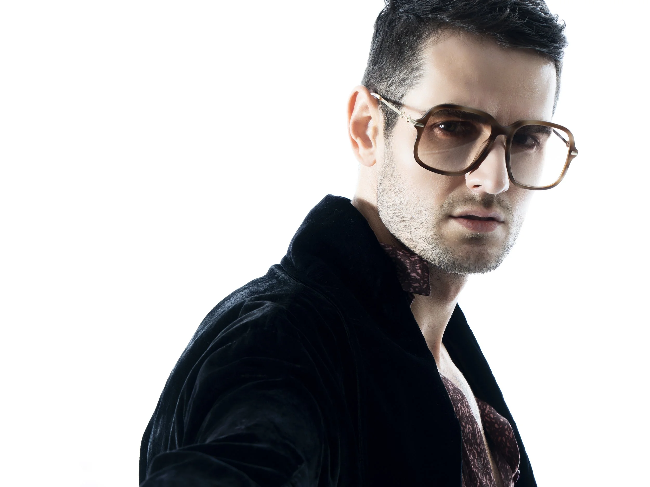 A confident man with dark hair, wearing large glasses, a black velvet jacket, and a patterned shirt, looking over his shoulder against a white background.