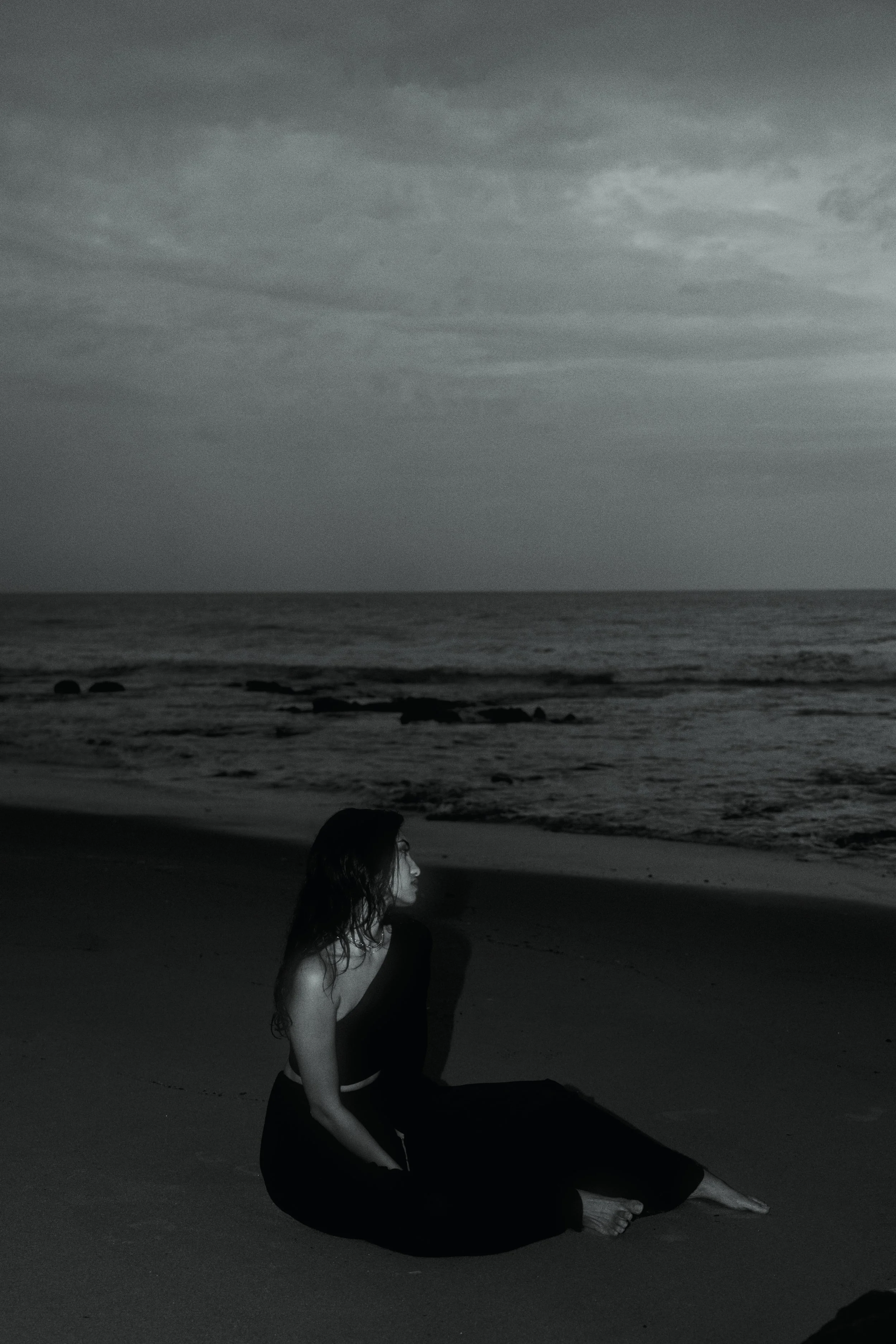 A woman with dark hair sitting on the sand near the ocean at dusk, looking towards the horizon over the water under a cloudy sky.