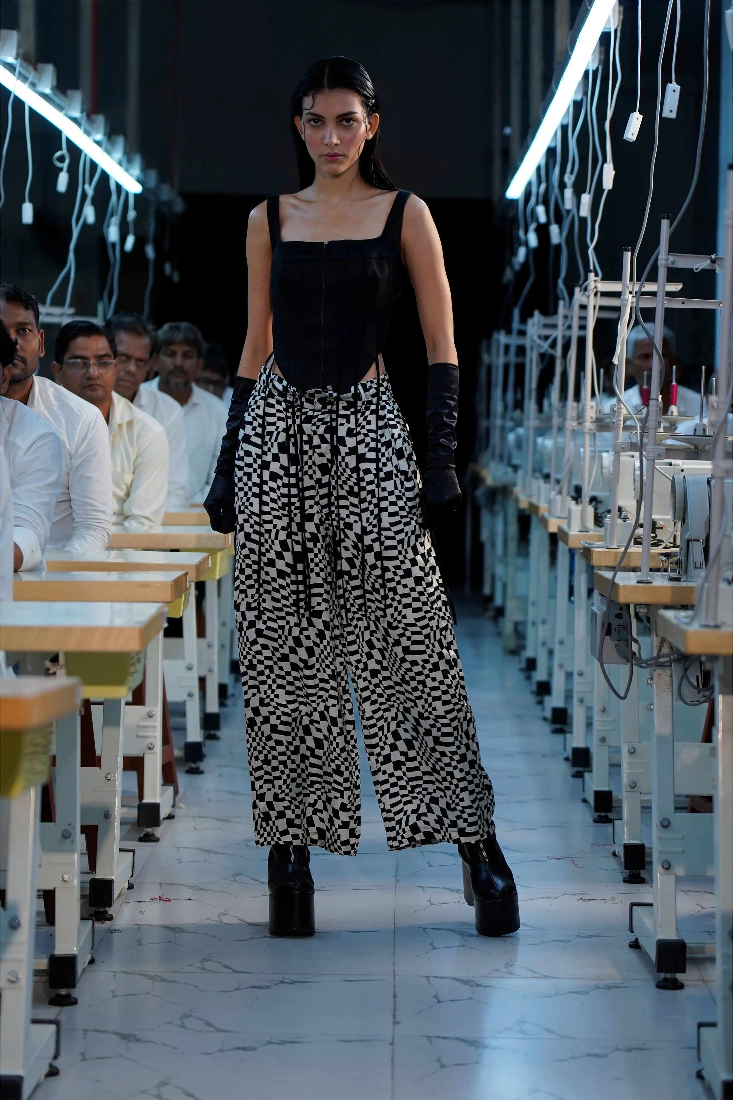 A female model walks down a runway in a fashion show, wearing a black top, checkered wide-leg pants, and platform shoes. Audience members are seated on both sides of the runway, and sewing machines are visible along the sides.