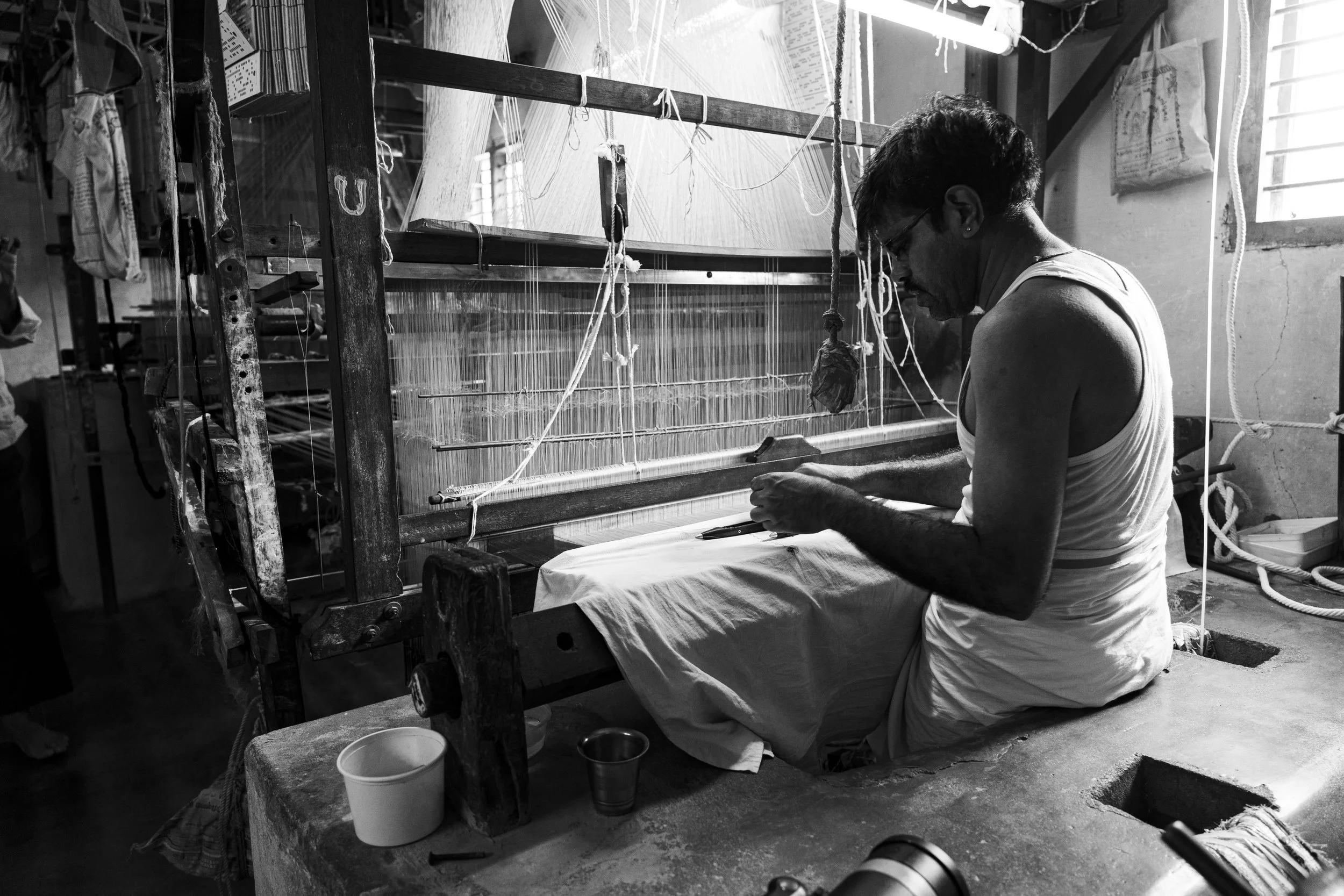 A man sitting at a loom, working on weaving fabric in a workshop with natural light coming through a window.