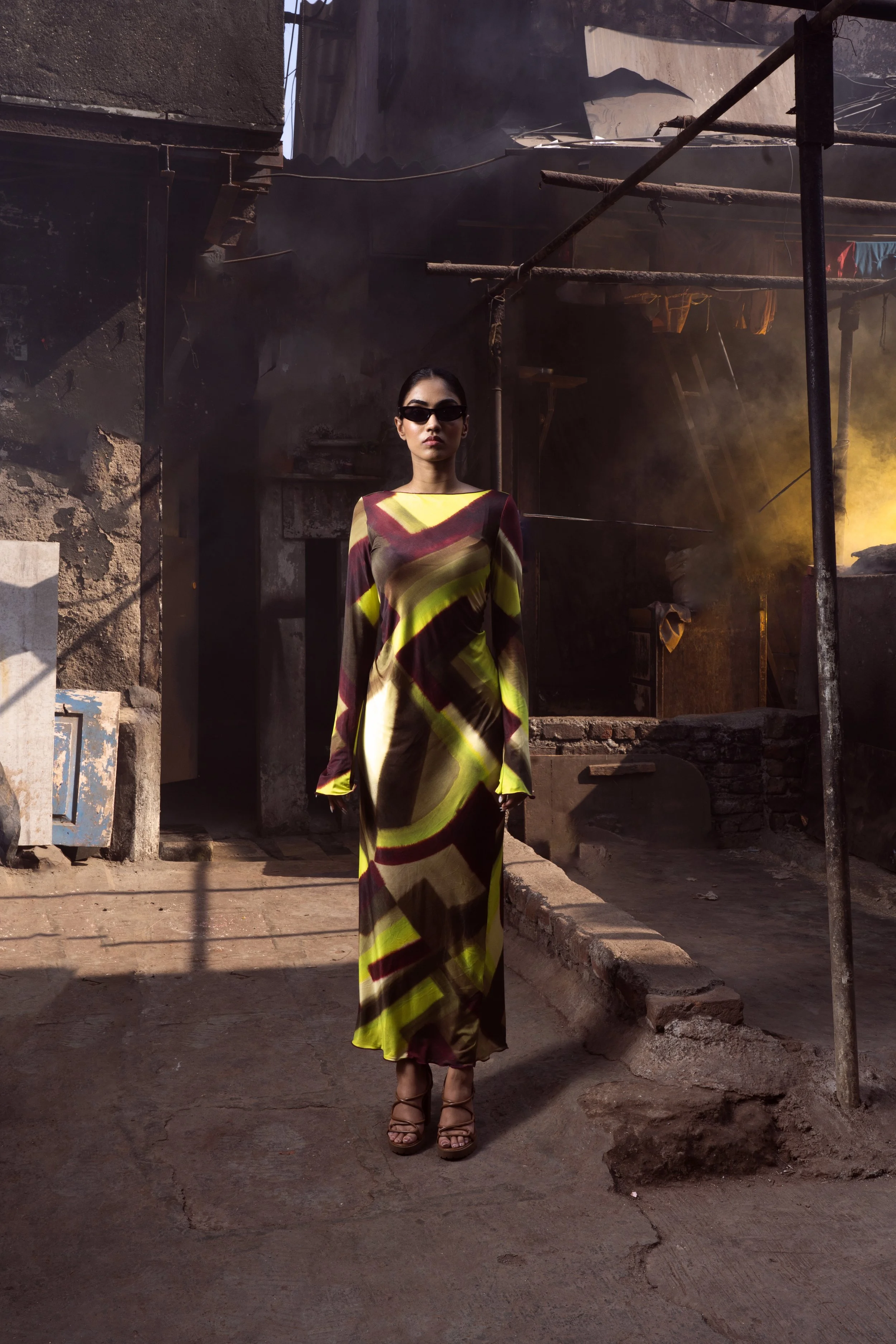 A woman in sunglasses and a colorful, patterned long dress stands in an urban, industrial setting with smoke and yellow light in the background.