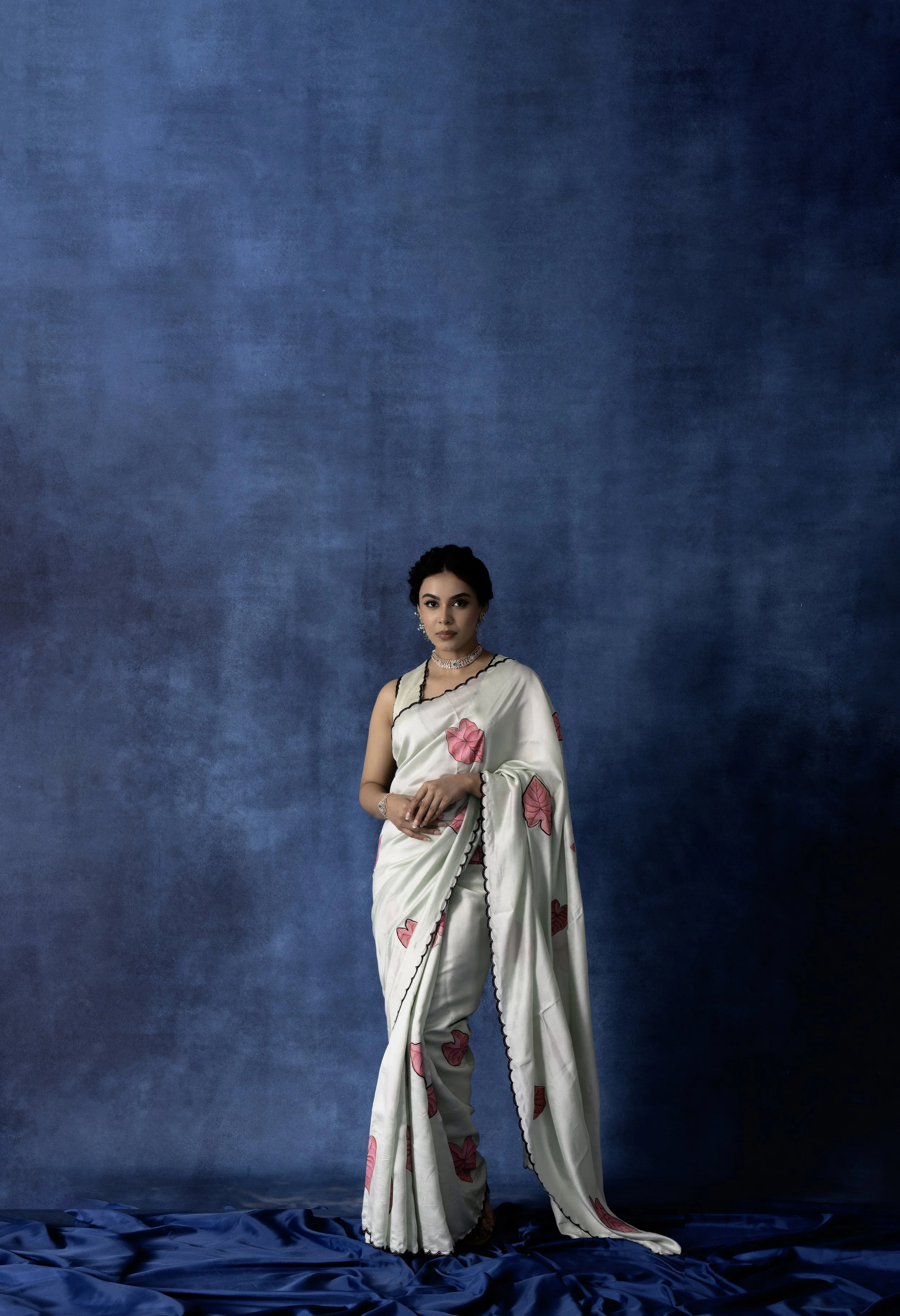 A woman wearing a cream-colored sari with pink butterfly patterns and black scalloped edges, standing against a textured blue background.