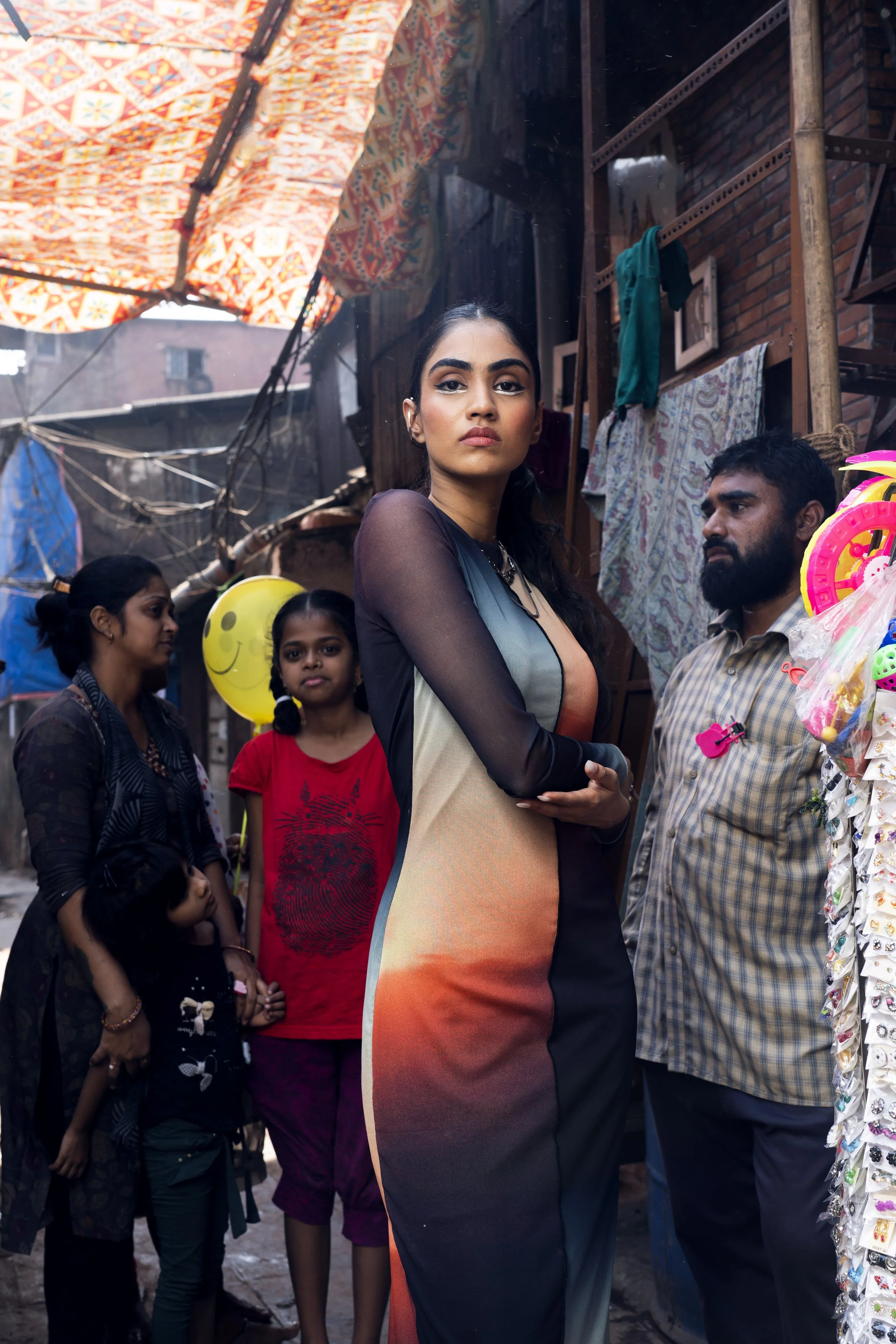 A group of people standing in a narrow alley with colorful umbrellas overhead and clothing hanging on the walls. The central woman is looking confidently at the camera with her arms crossed.