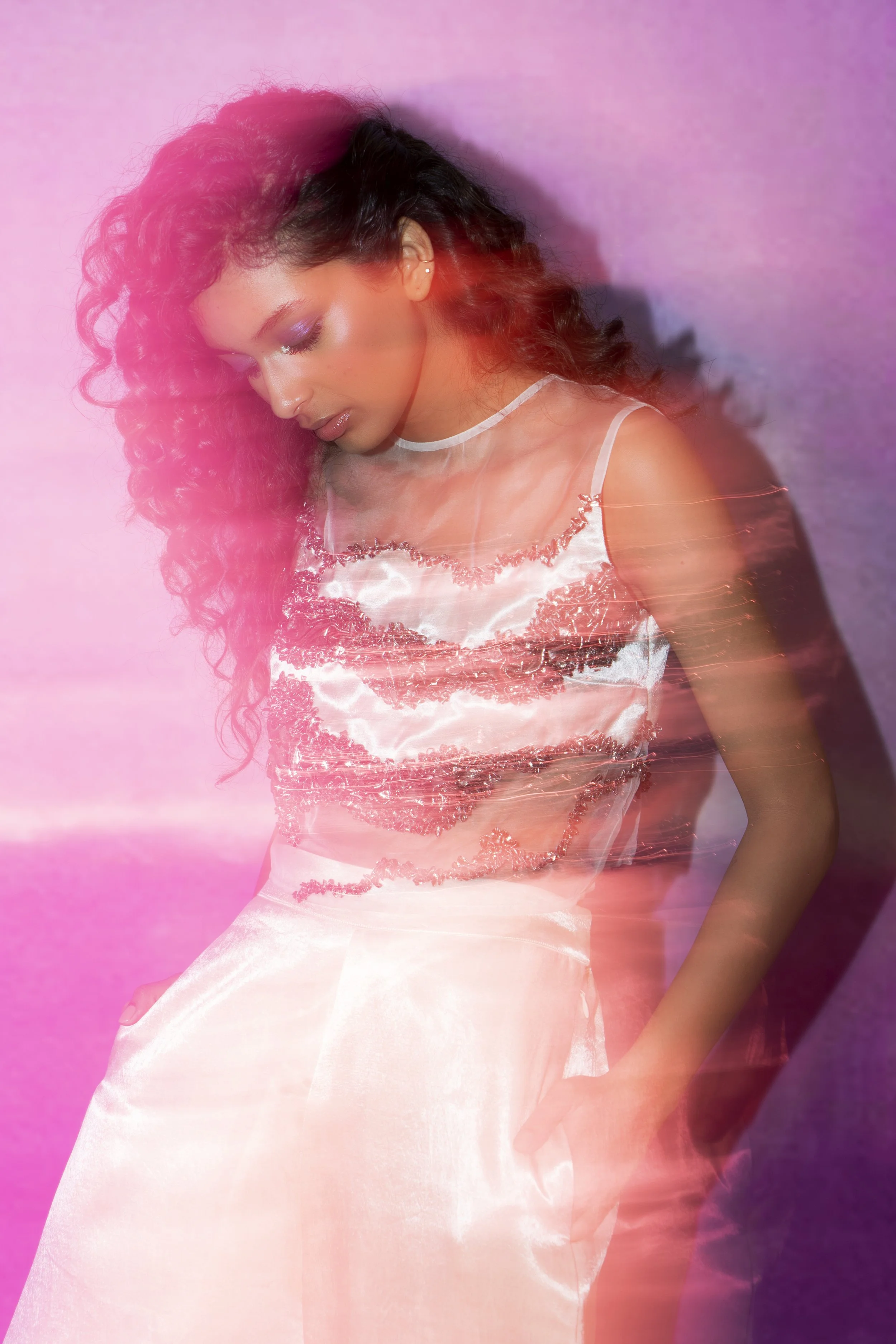 A woman with long curly hair wearing a shiny, embellished top and a light-colored skirt, standing against a pink and purple background with a pink light streak across her face and body.