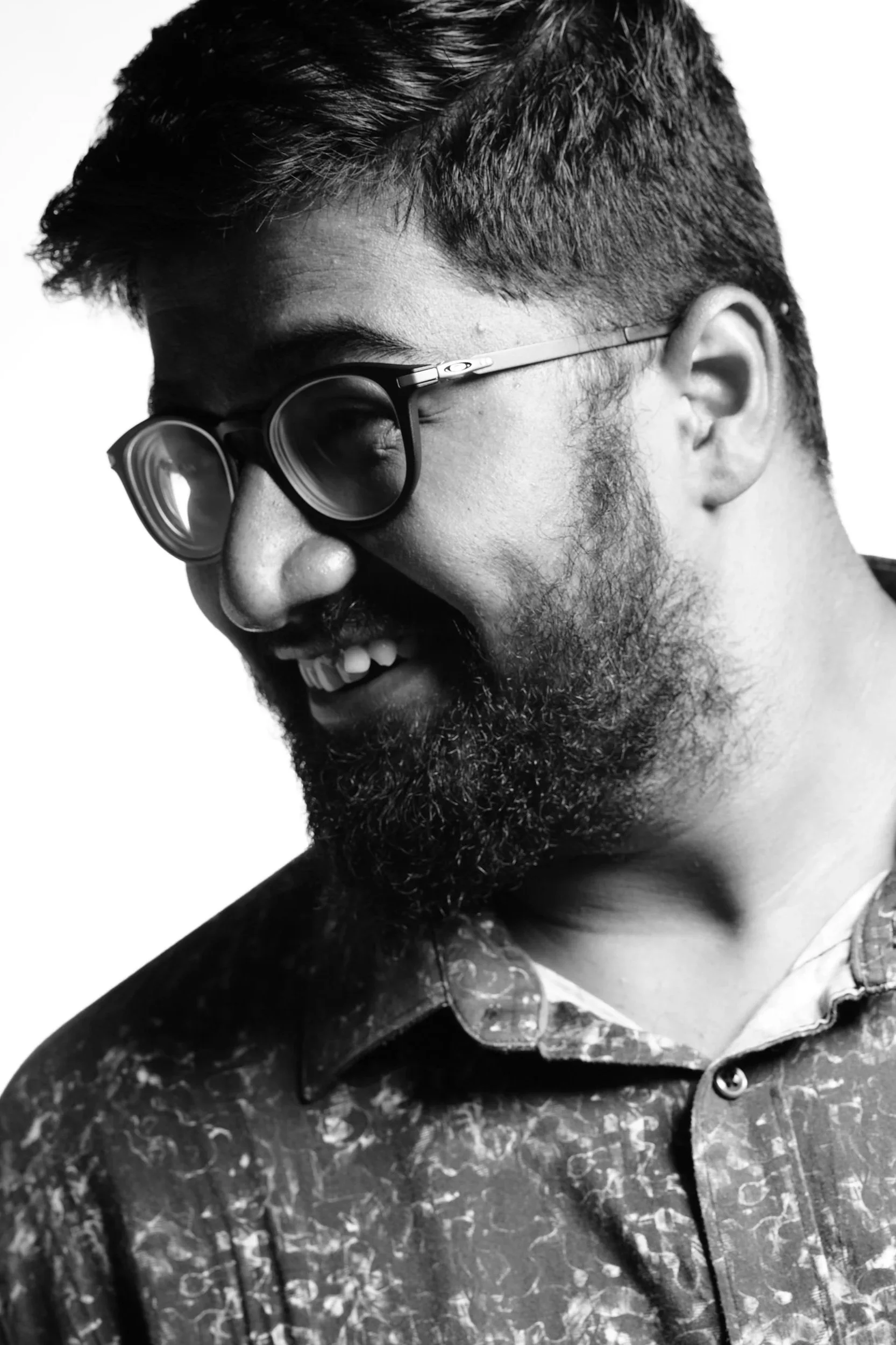 A black and white close-up portrait of a man with short, dark hair, a beard, glasses, and a patterned jacket, smiling gently with his eyes closed. Portrait of the ,man himself