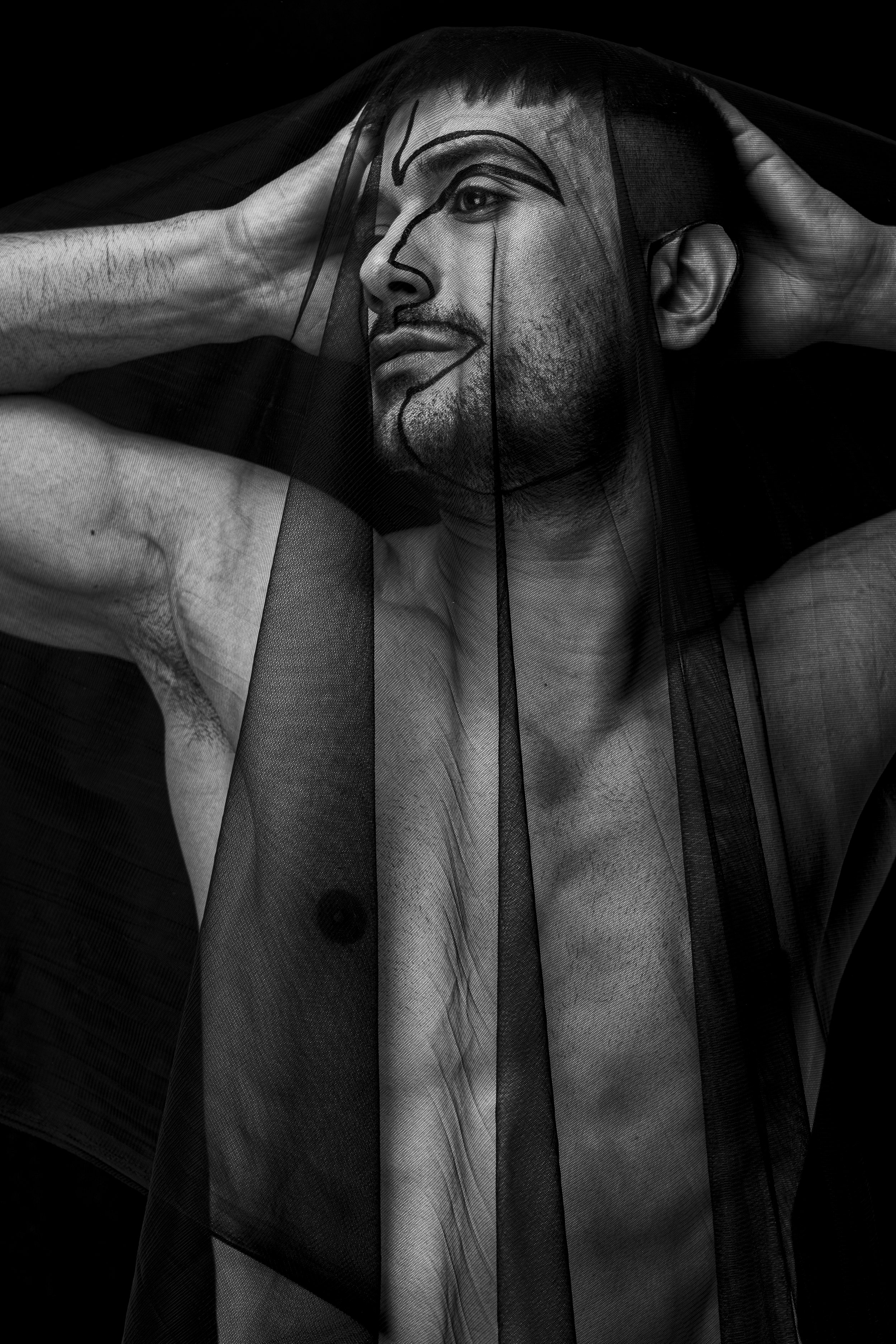 Black and white photograph of a man with tattoos on his arms, partially covered by a sheer black fabric with artistic face makeup underneath.