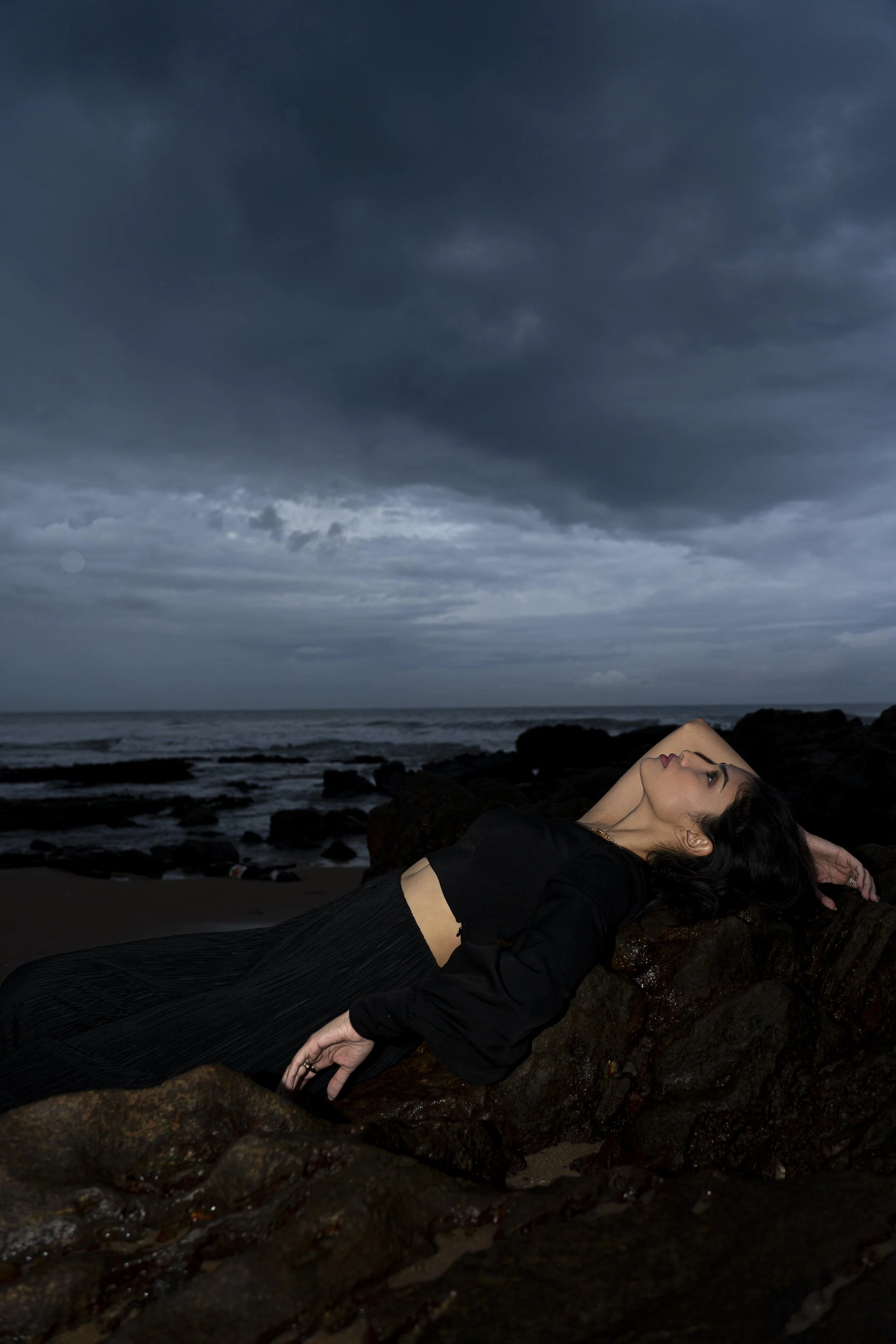 A woman with dark hair lying on rocks at a beach during stormy weather, with dark clouds overhead.