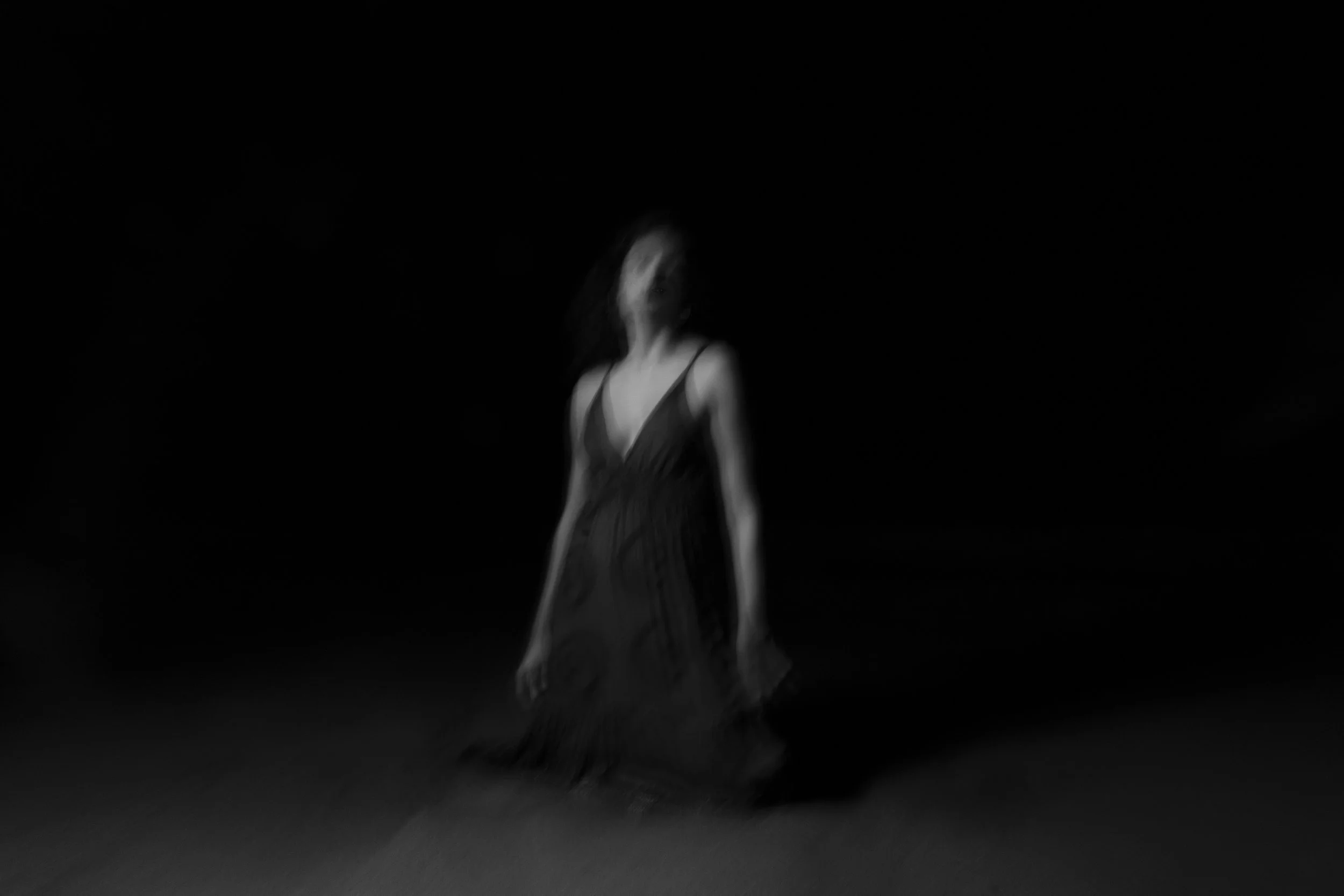 Blurry black-and-white image of a woman in a dress sitting or kneeling on the ground in a dark setting.