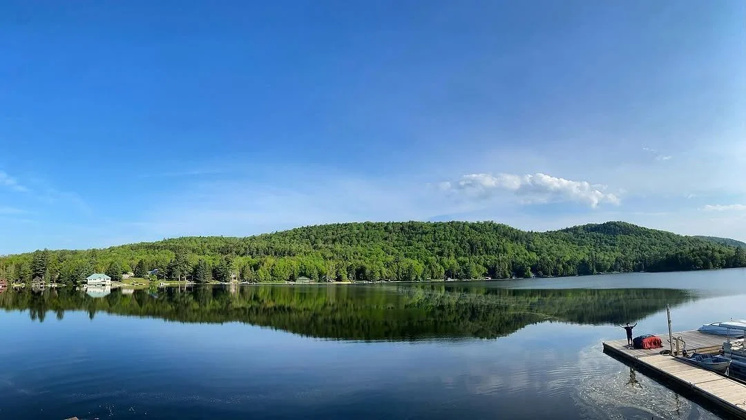 A calm lake with a clear reflection of a green forested hill under a blue sky with a few clouds, and a dock with a person standing at the end, near boats.