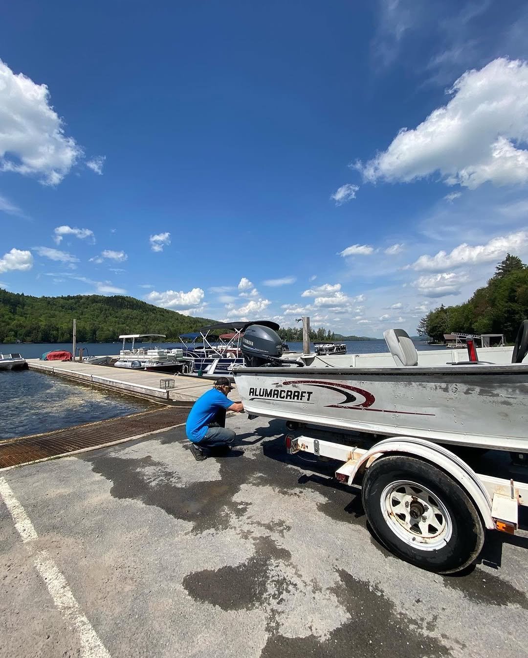 A person in a blue shirt and dark pants crouches next to a boat on a trailer at a lakeside dock on a bright, sunny day. The boat has the brand name 'Alumacraft' on its side and an outboard motor attached.
