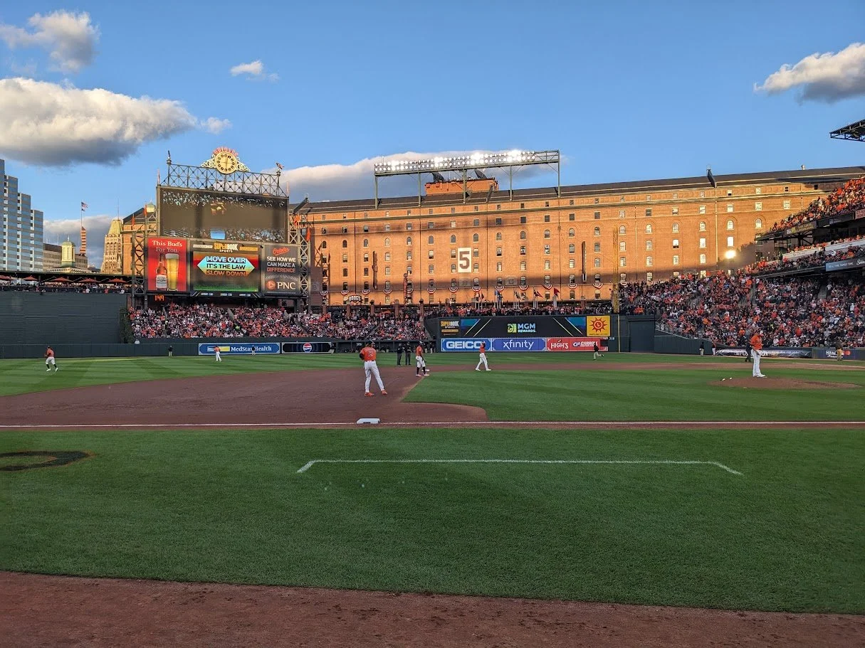 A baseball game in progress at a stadium filled with spectators. Players are on the field with a cityscape in the background.