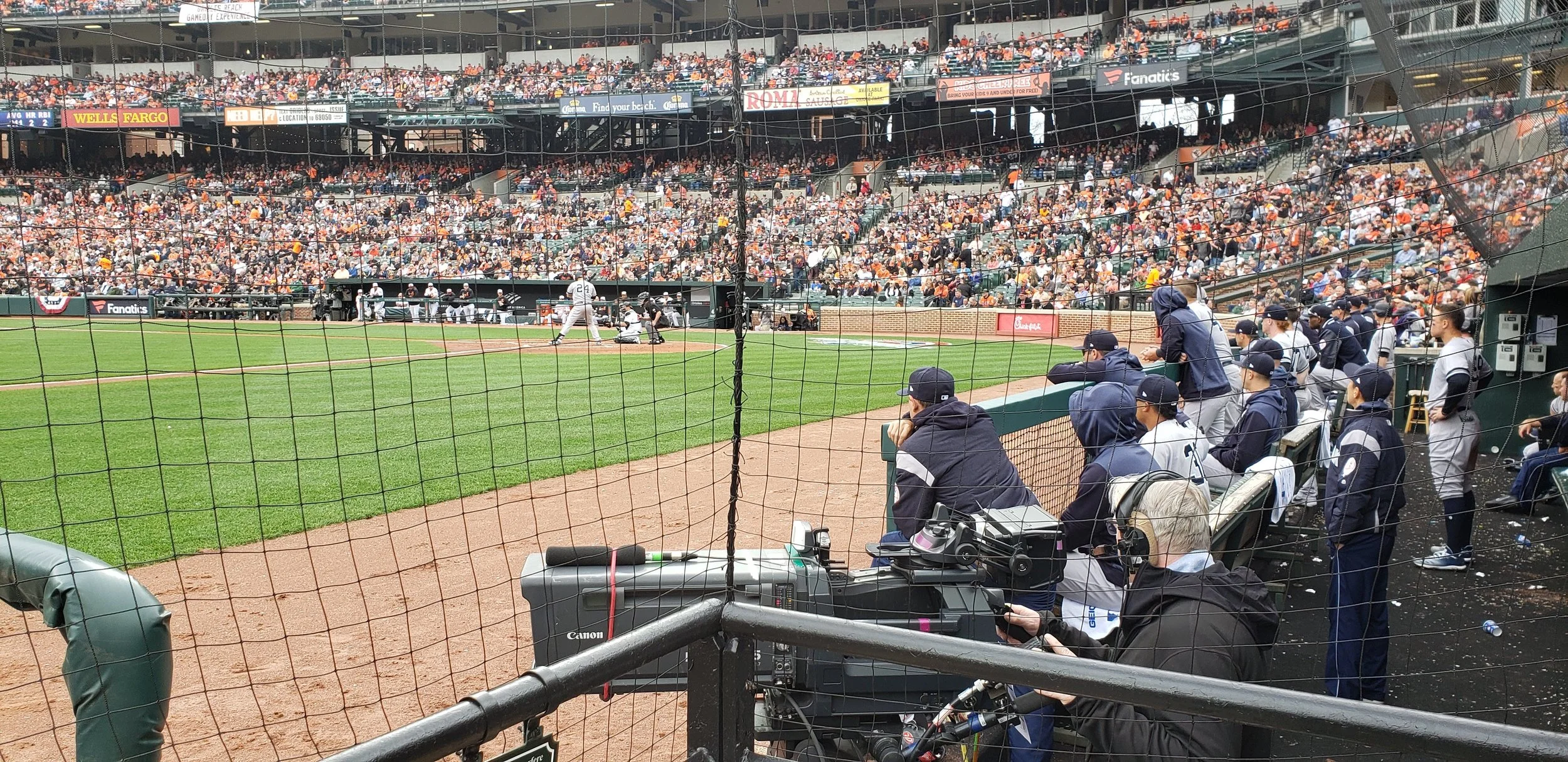 A baseball game in progress at a stadium, with players on the field and a crowd in the stands. The view is from the dugout area with several coaches and team members watching the game.