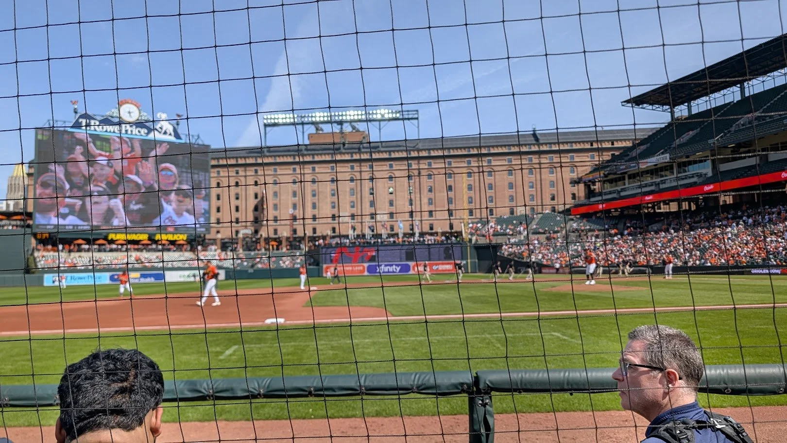 View of a baseball stadium from behind home plate, showing players on the field, a large video screen with spectators, crowd in the stands, and a building in the background, with a clear blue sky.