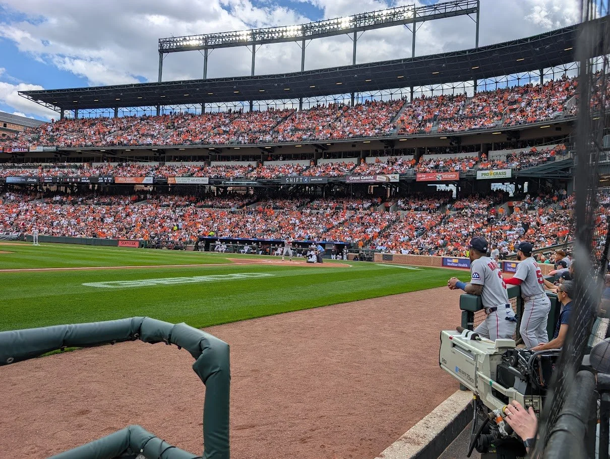 A baseball game in progress at a stadium with a large crowd, players on the field, and camera operators near the dugout.