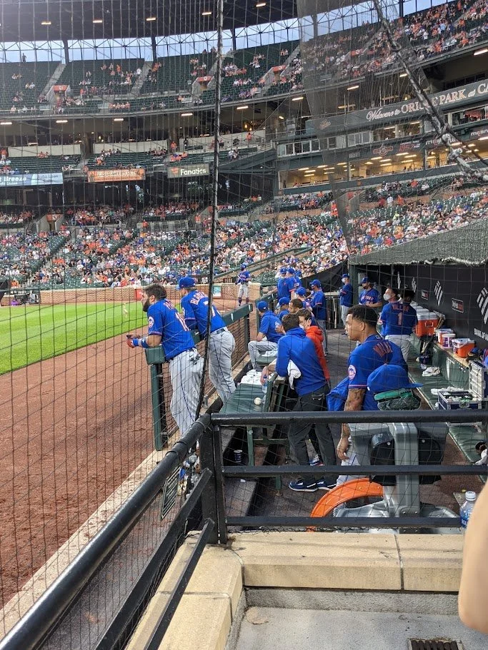 Baseball players in blue uniforms in the dugout at a stadium during a game. The stadium is filled with spectators, and the field is visible beyond the dugout.