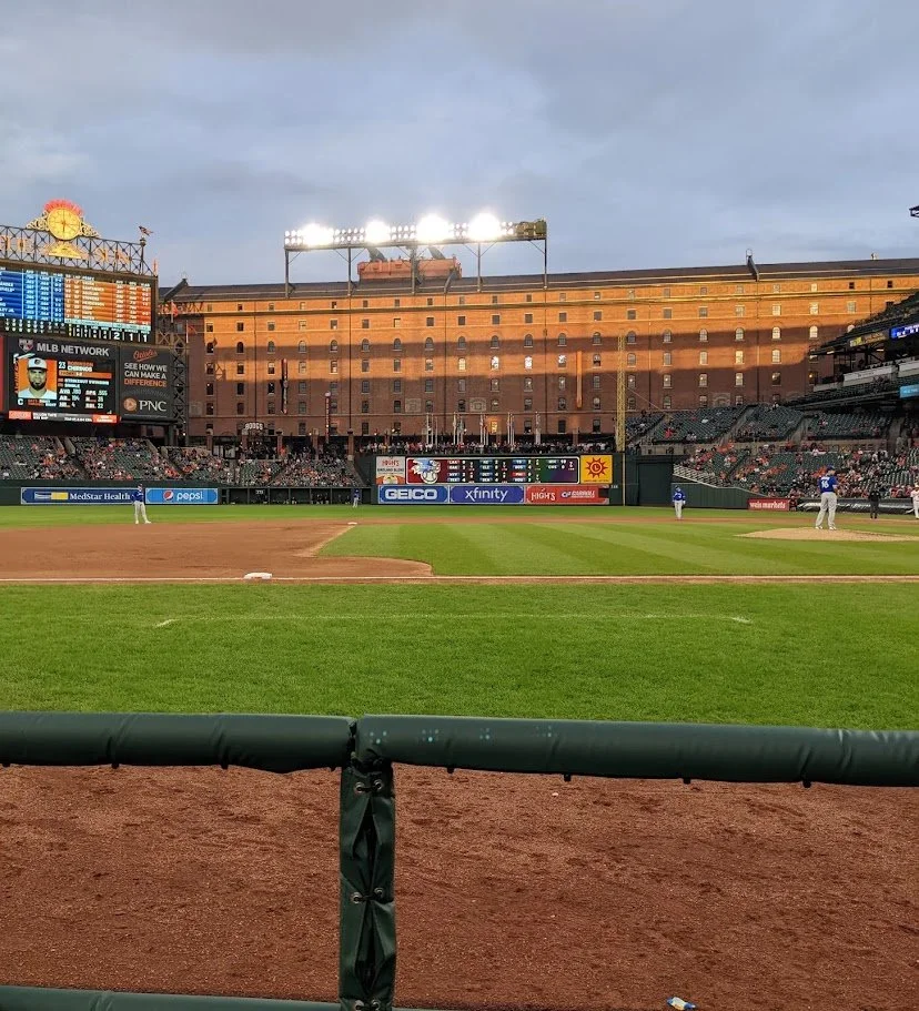 View of a baseball stadium with a grassy field, players on the field, scoreboard, and advertising signs, taken from behind the protective railing.