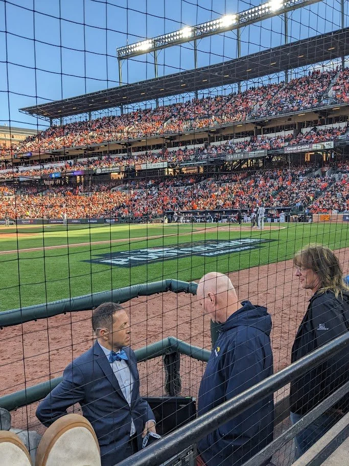 People standing near the railing at a baseball stadium with a crowd in the background, visible through a protective net.
