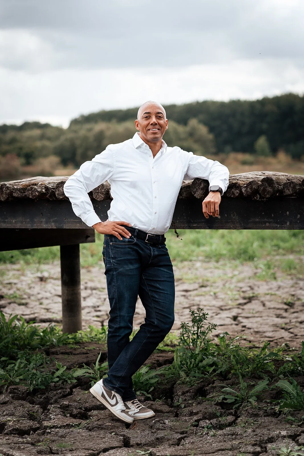 Man in witte blouse en jeans leunt tegen een houten reling buiten, groene heuvels en een bewolkte hemel op de achtergrond.