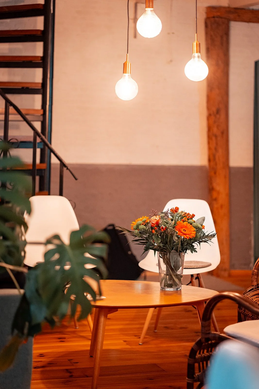 Een gezellige woonkamer met een houten tafel, een vaas met oranje bloemen, witte stoelen, hangende lichtbulben en een trap op de achtergrond.