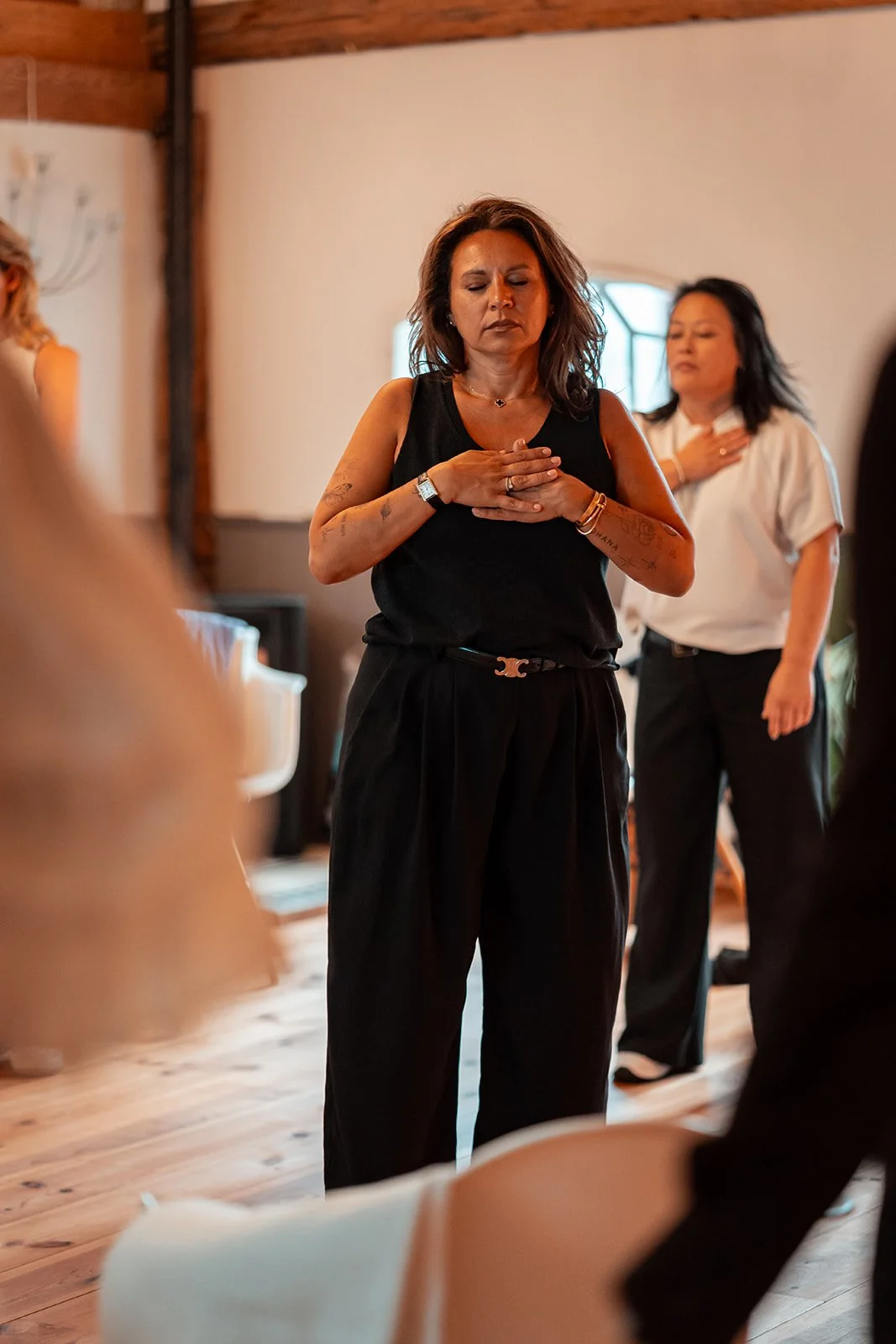 Vrouw met gesloten ogen en handen op haar borst in een rustige houding tijdens een spirituele of meditatieve oefening, omringd door andere mensen in een lichte, gezellige ruimte.