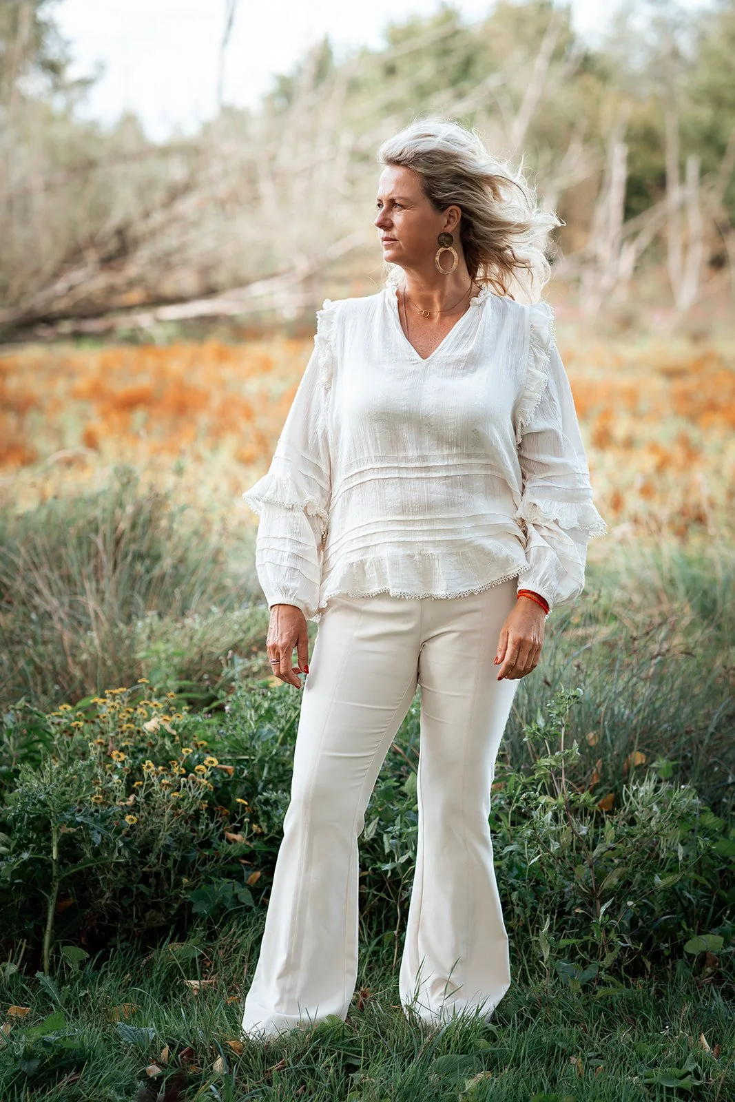 Een vrouw met grijs haar die in een natuurlijke buitenomgeving staat, gekleed in een witte blouse en witte broek.