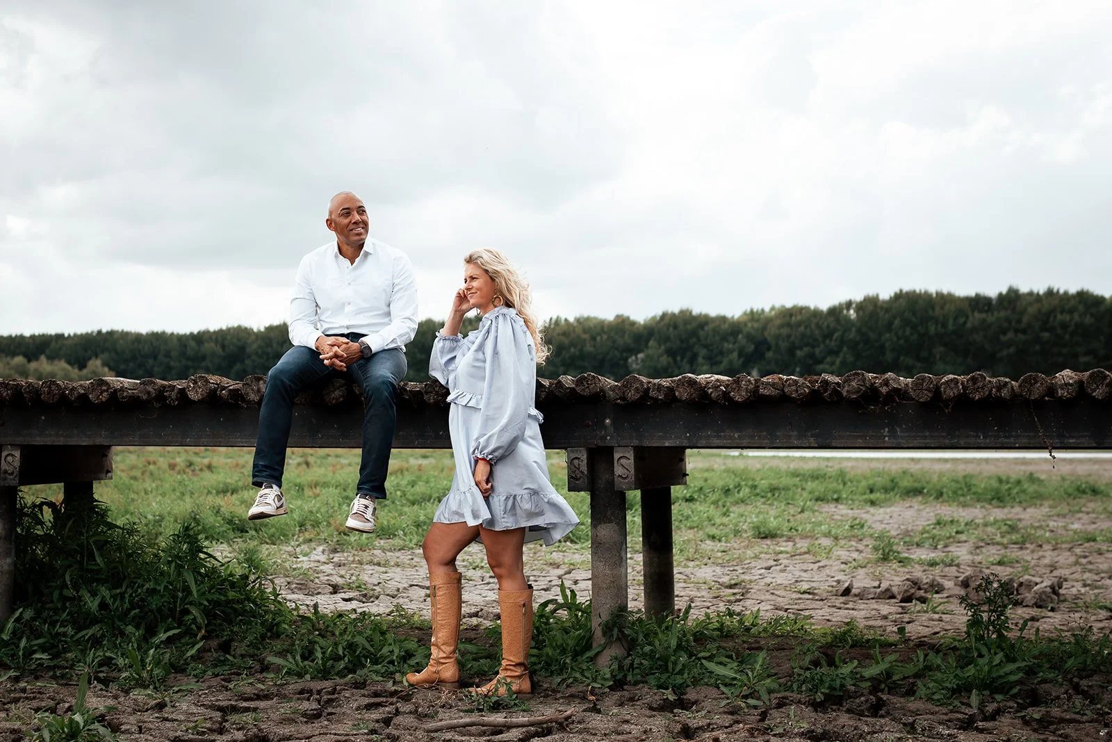 Twee mensen, een man en een vrouw, zitten en staan bij een houten bankje in een open veld met bomen op de achtergrond en een bewolkte lucht.