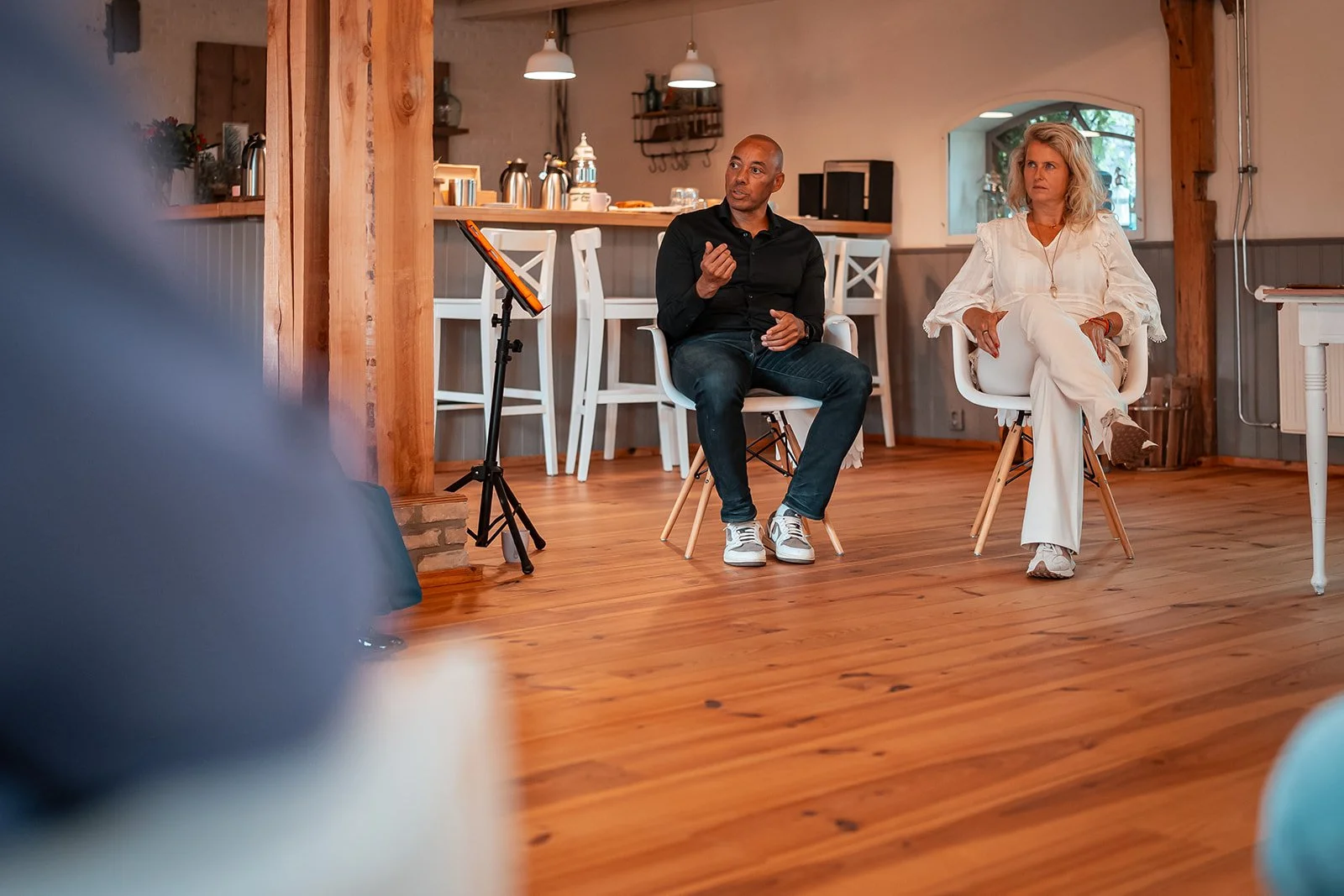 Twee mensen zitten in een huiselijke ruimte, mogelijk tijdens een gesprek of interview. De man links is van middelbare leeftijd, heeft een bald head en draagt een zwarte shirt en jeans. De vrouw rechts heeft blond, golvend haar en draagt een witte outfit. Ze zitten op witte stoelen op een houten vloer, met een achtergrond van een keuken met witte barstoelen en voorwerpen.