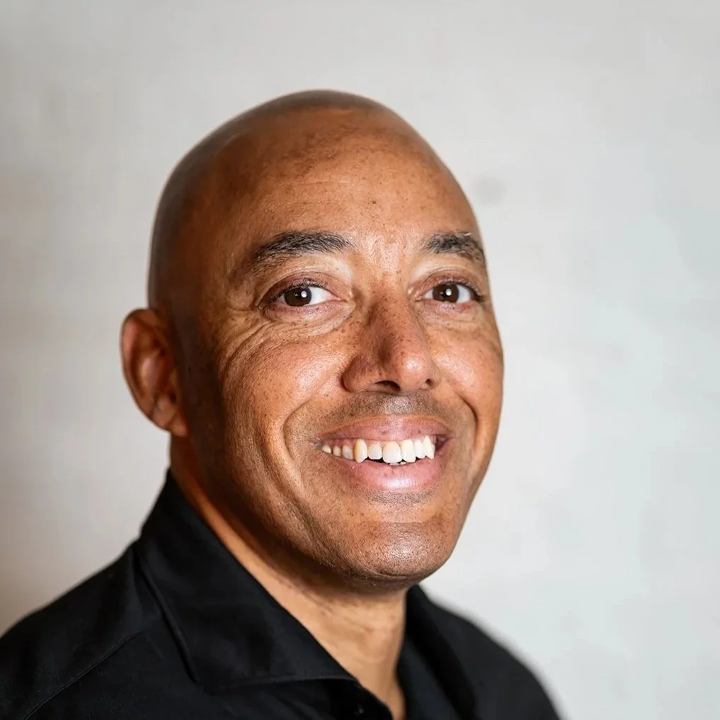 Portret van een lachende man met een kaal hoofd en een zwarte blouse, voor een lichte achtergrond.