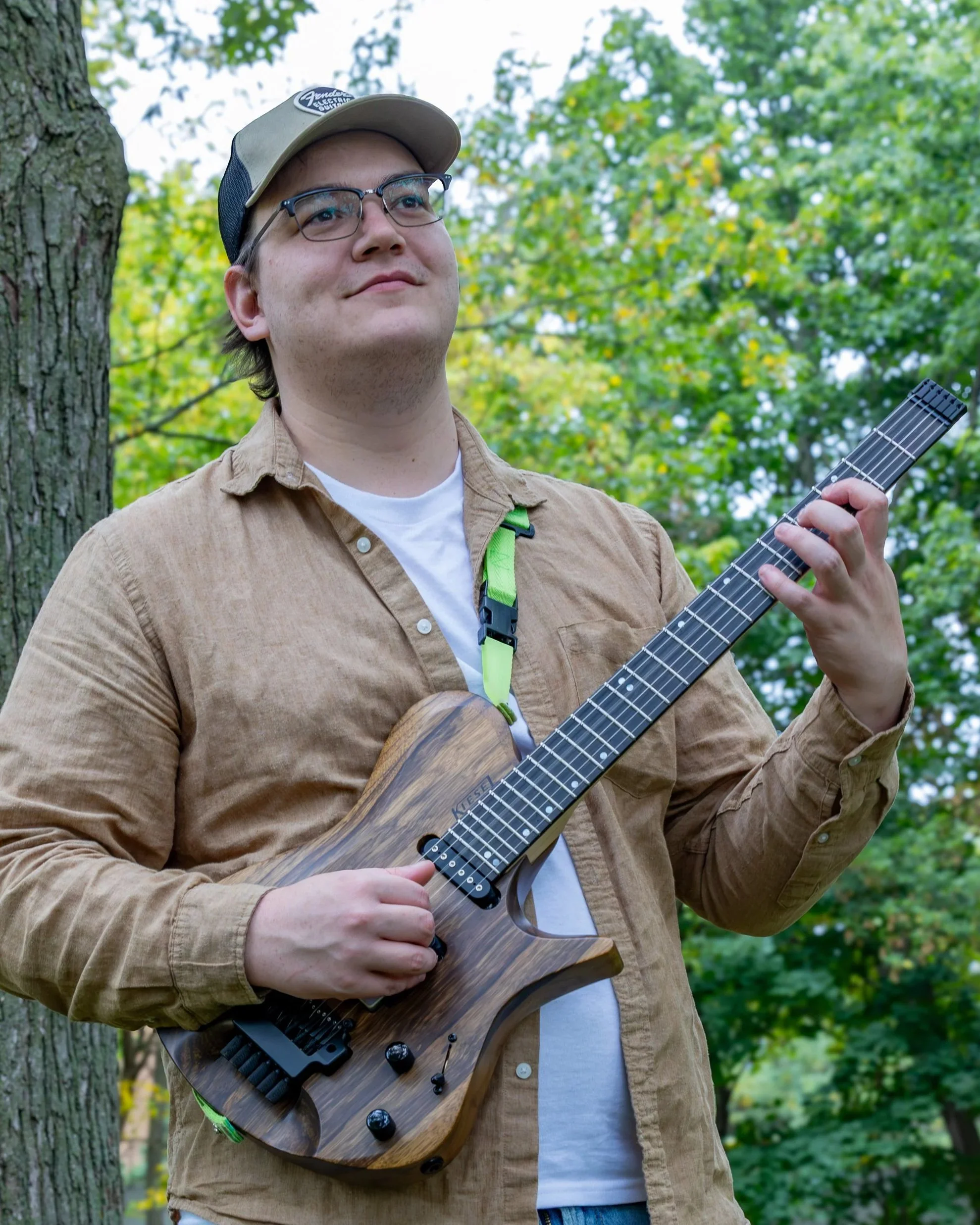 A young man with glasses and a beige cap playing an electric guitar outdoors in a wooded area.