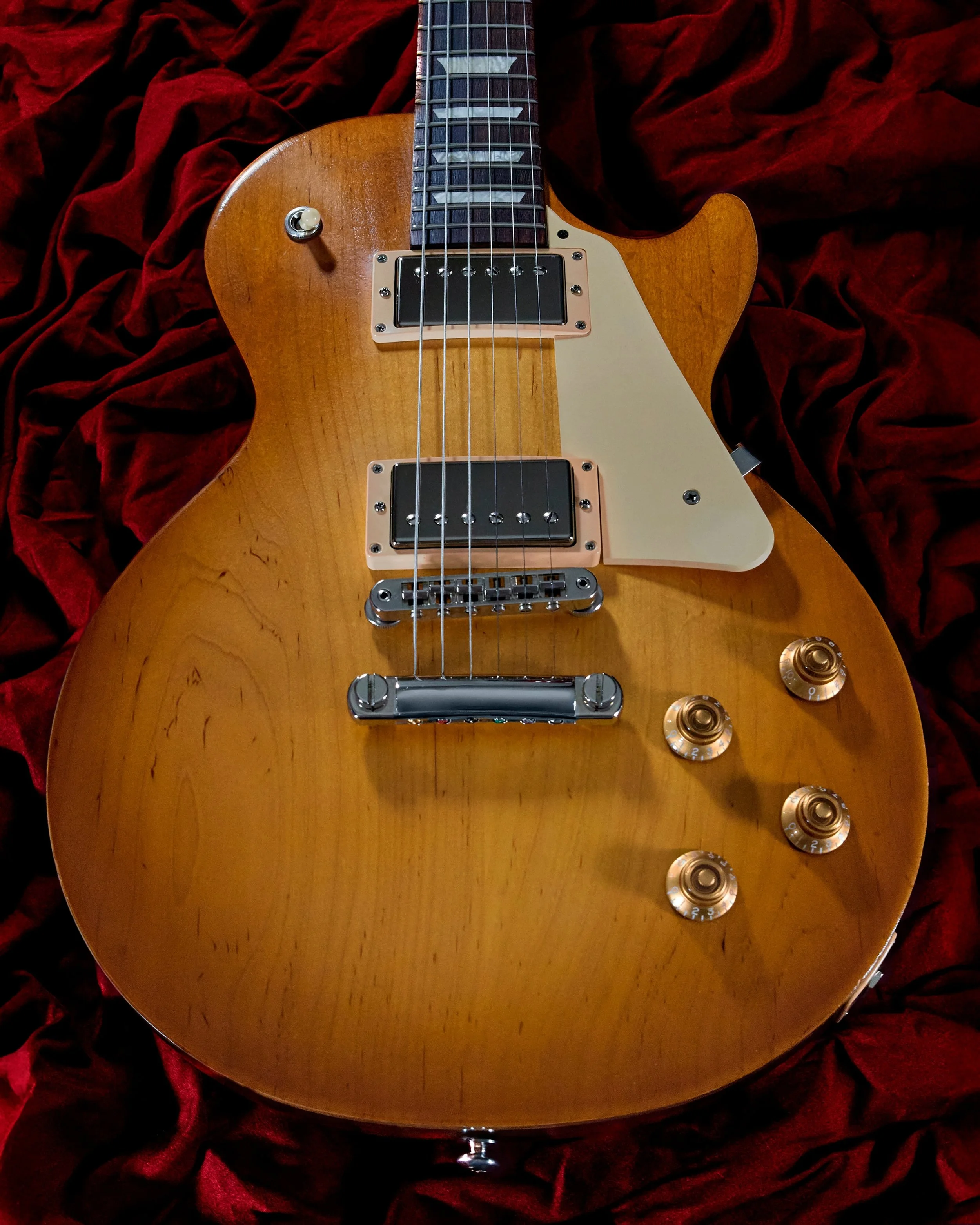 A vintage electric guitar with a natural wood finish, four control knobs, two pickups, and a white pickguard, resting on red velvet fabric.