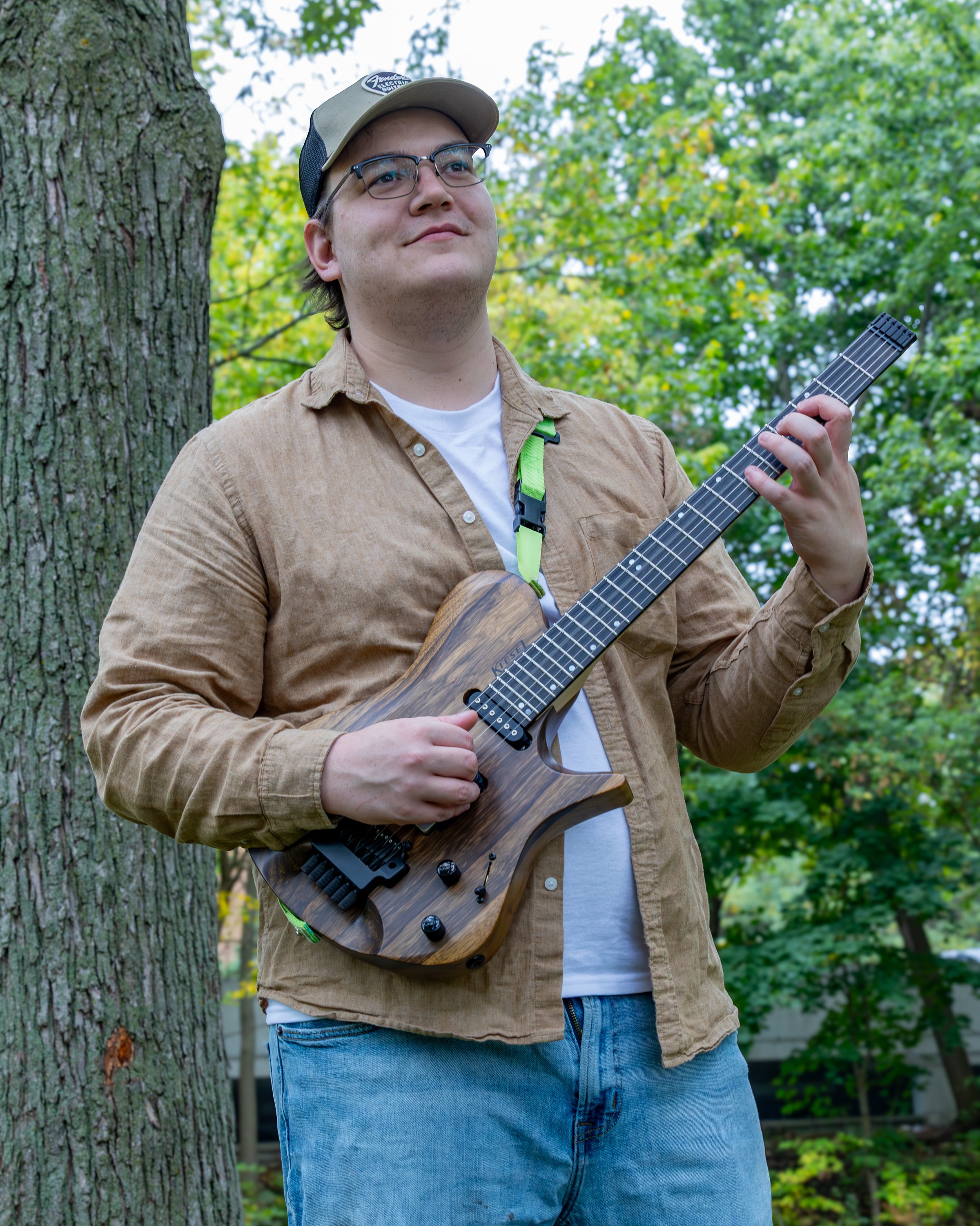Young man outdoors, wearing glasses and a beige cap, playing a brown electric guitar near a tree with green leaves in the background.