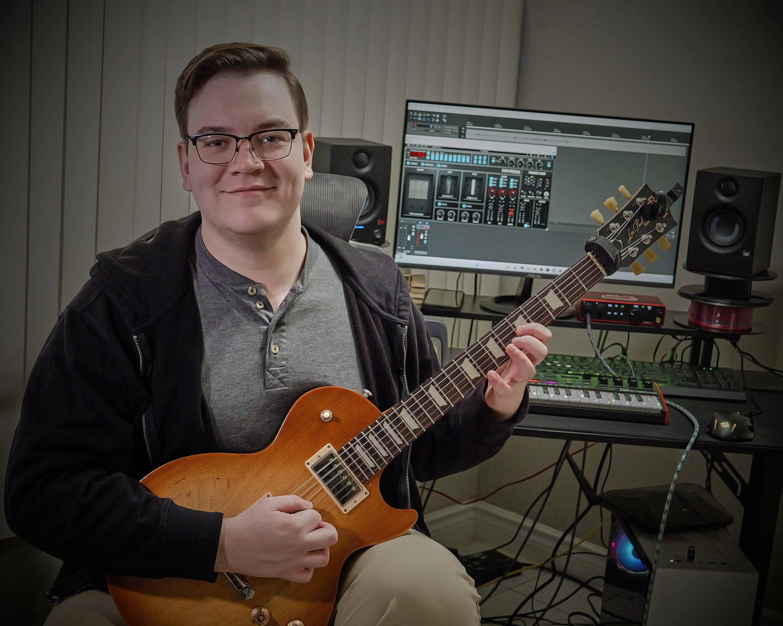 A man in glasses sitting in a music studio holding an electric guitar, with a computer and audio equipment visible on his desk behind him.