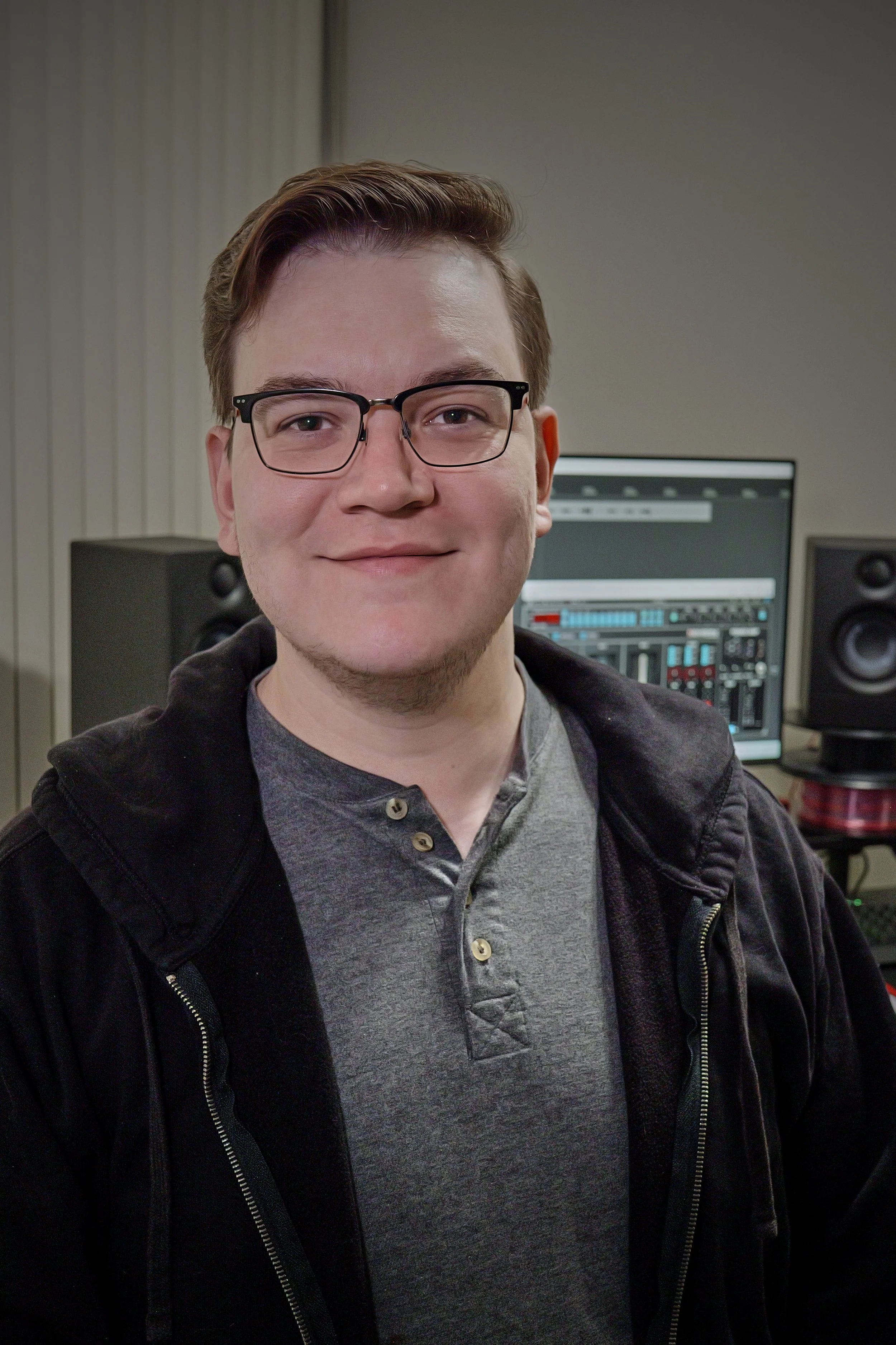 A young man with glasses, wearing a gray shirt and black hoodie, smiling in a room with audio equipment and a computer monitor in the background.
