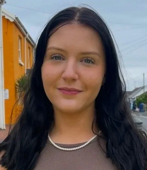 A young woman with long black hair and fair skin smiling outdoors, wearing a pearl necklace and a sleeveless top, with a yellow building and overcast sky in the background.