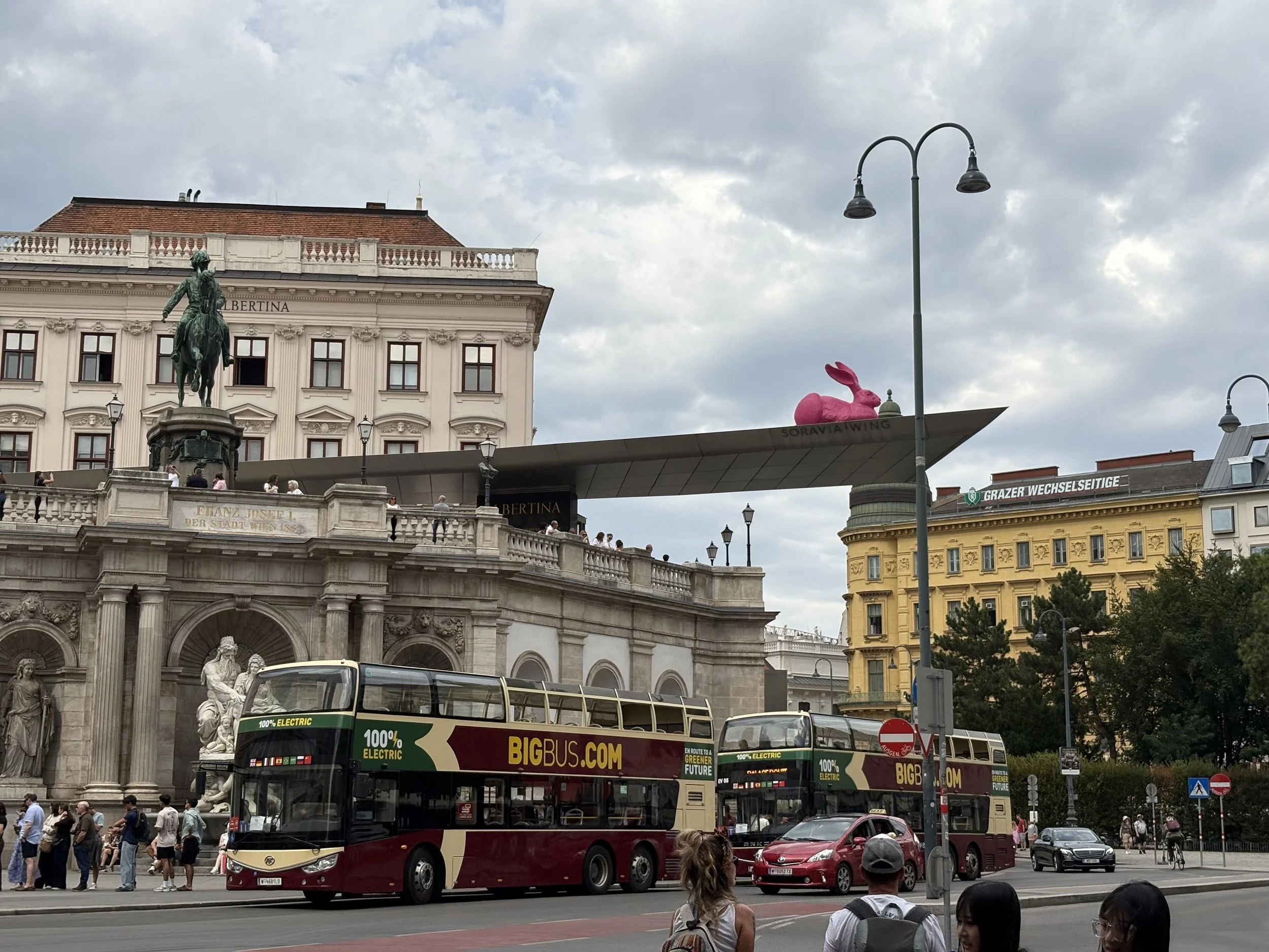 Photo of an ornate stone building with columns and statues and arches partially blocked by two doubledecker buses. There is a viewing platform on the top of the building with an equestrian statue, and a long roof with a pink hare sculpture