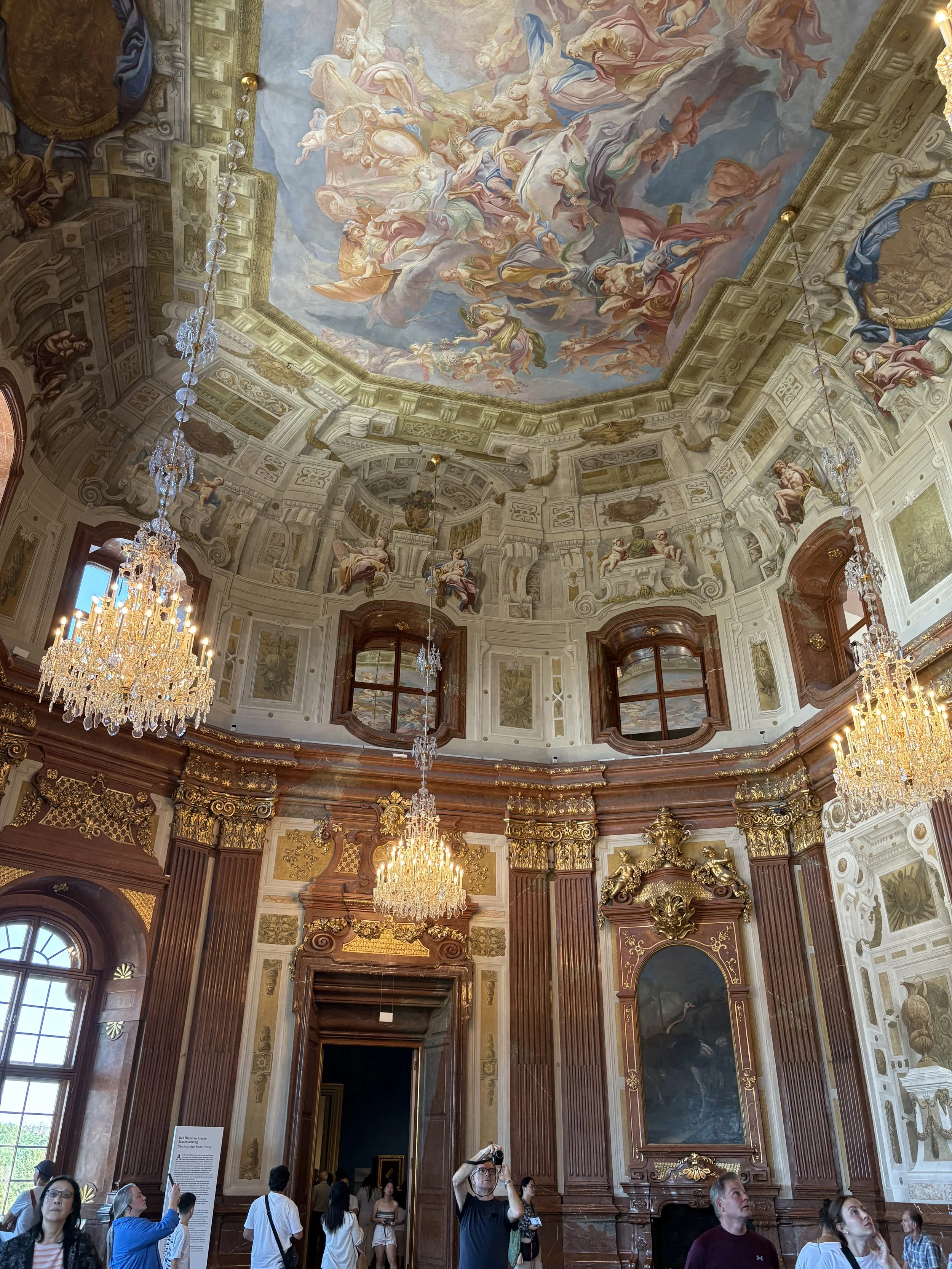 A room filled with tourists with an elaborately painted ceiling and very ornate brown and gold walls and crystal chandeliers