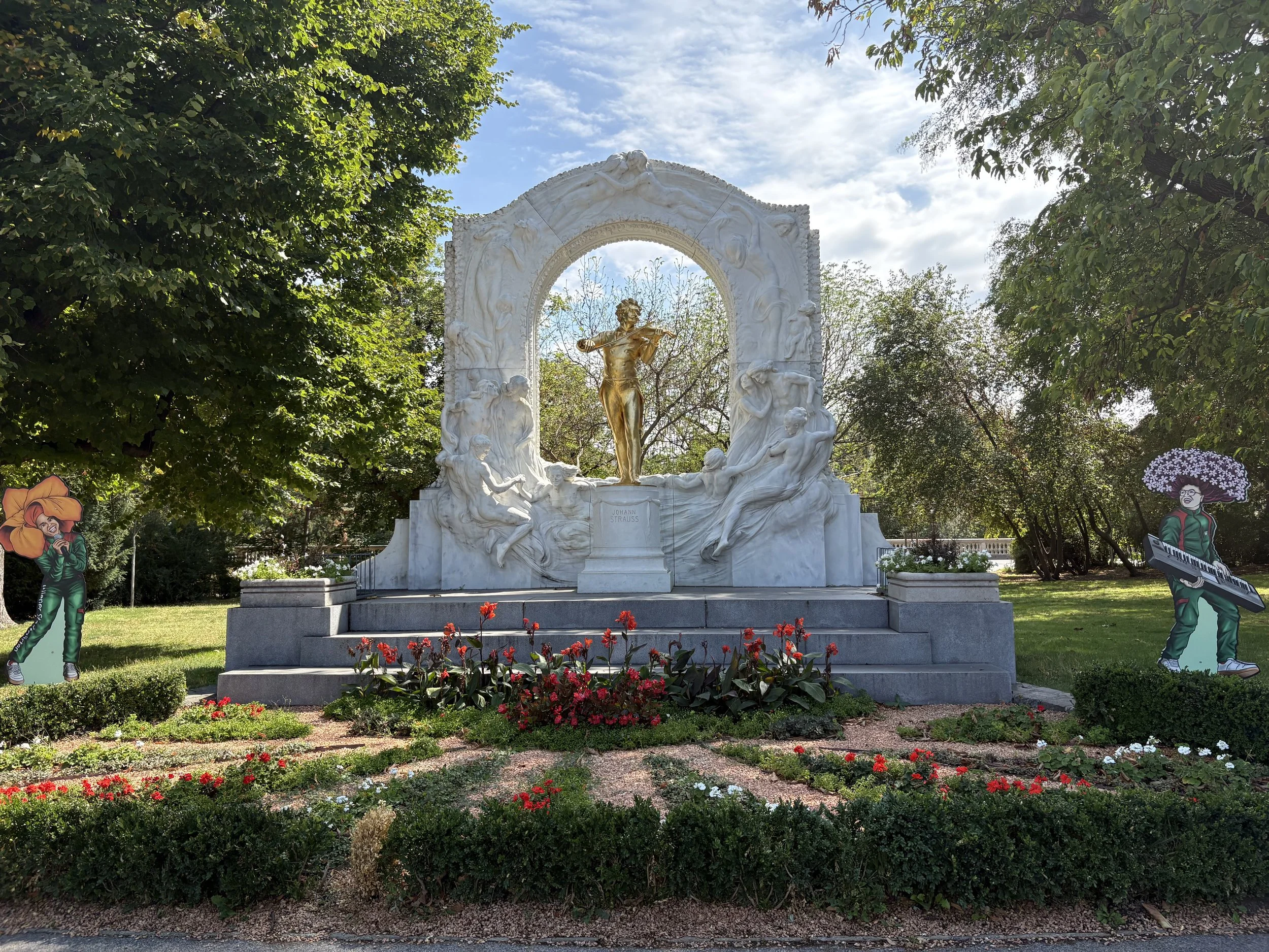 A gold statue of a man playing a violin surrounded by a sculpted white marble arch. There are red flowers in front and trees around the statues