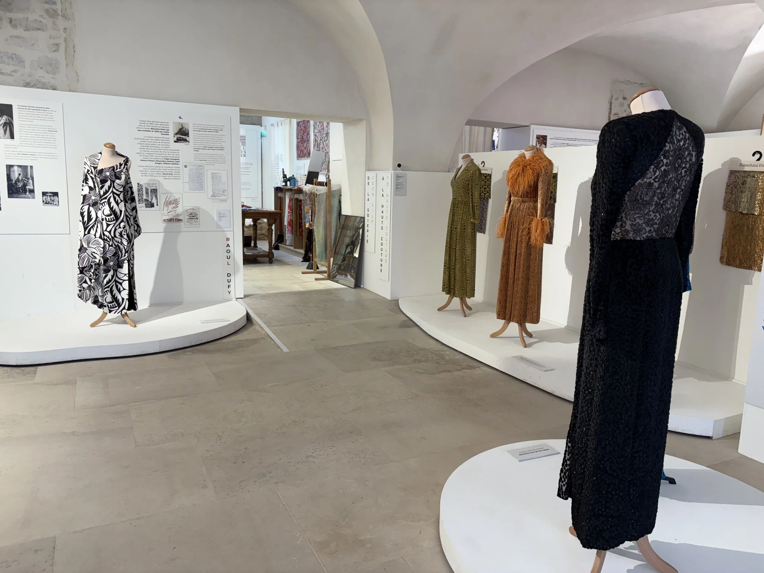 Photo of the inside of a museum. There are several gowns: a black and white design with long sleeves, an olive green dress, a rust orange dress, and a black dress with long sleeves. The floor is grey stone and the walls are white and arched.