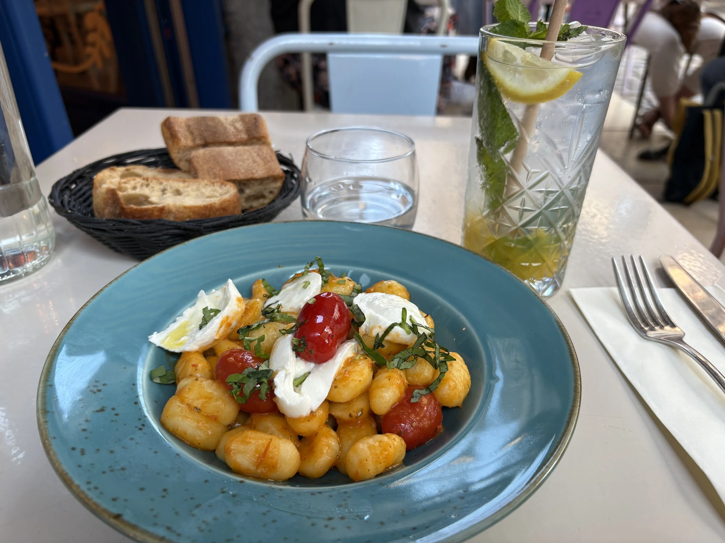 Photo of a table with a dish of gnocchi with mozzarella, shredded basil, and cherry tomatoes. There is a basket of sliced baguette and a short glass of water. There is a tall glass with orange puree in the bottom, mint, and a lemon wedge.
