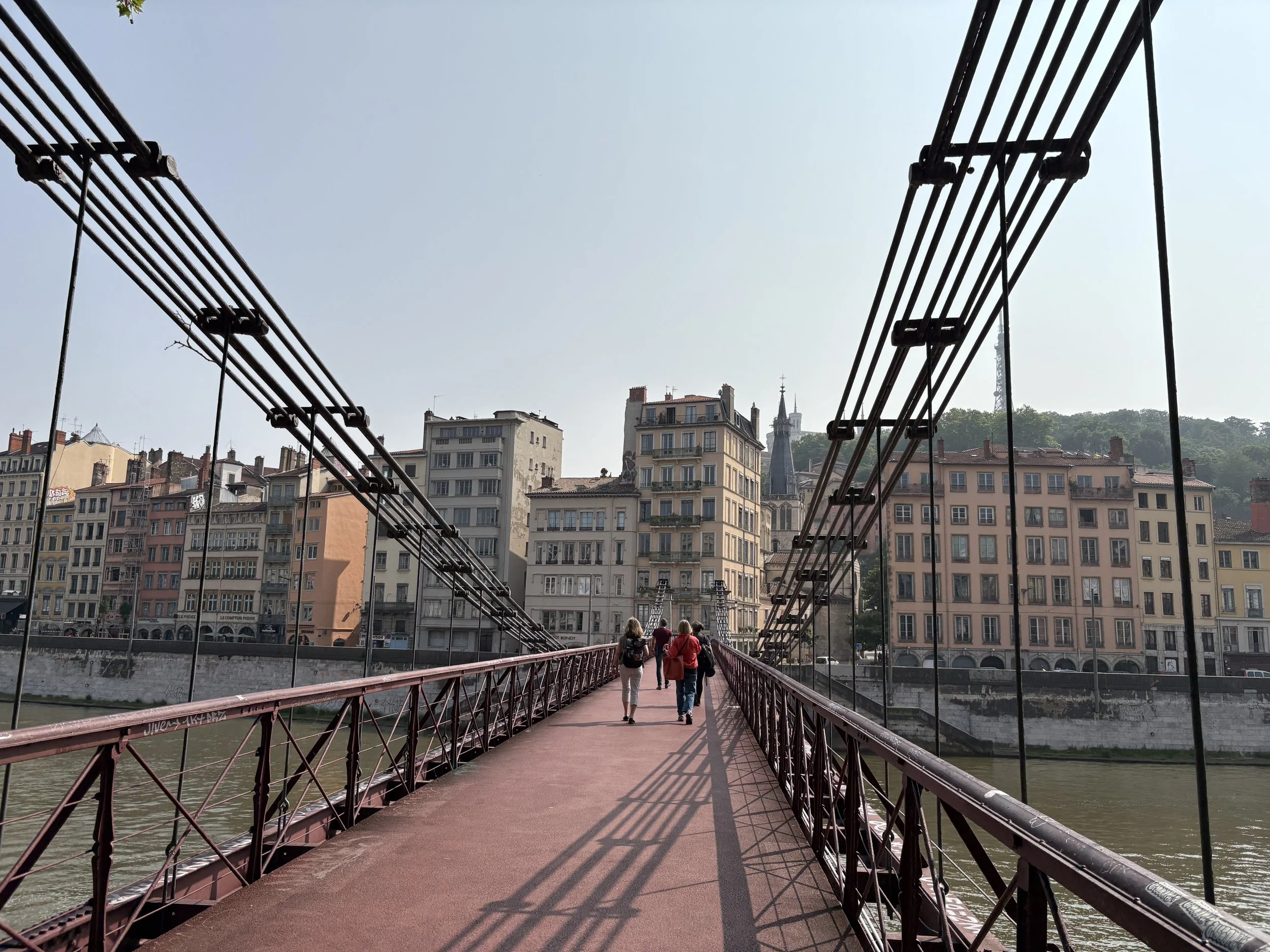 Photo of a red metal bridge leading straight back to a riverbank with tan and rust colored buildings. There are people crossing the bridge and a church spire in the distance.