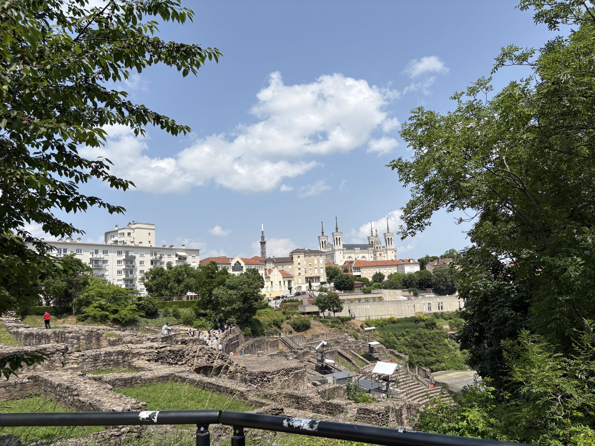Photo of stone ruins along a hill surrounded by bushes and trees. In the distance you can see a white building and four spires of a cathedral. The sky is blue with some white clouds.