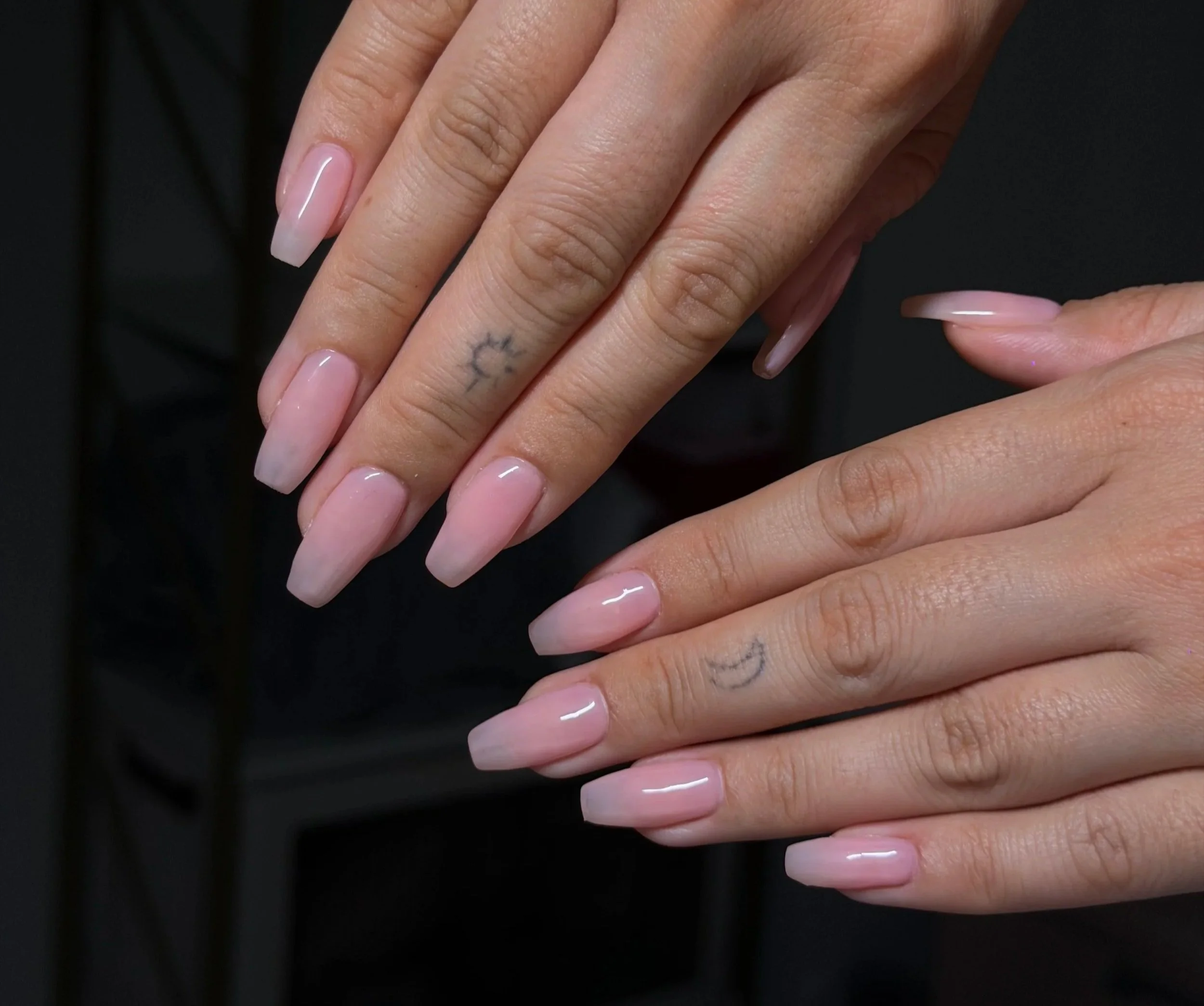 Close-up of a person's hands with long, well-manicured, French-style nails and a small tattoo of a crescent moon on one finger.