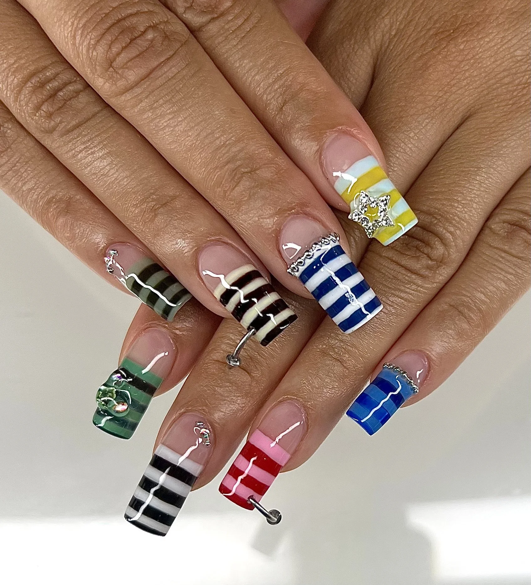 Close-up of nails with colorful striped nail art in red, pink, blue, green, yellow, white, black, and silver, with rhinestone accents and charm decorations, on a person's fingers.