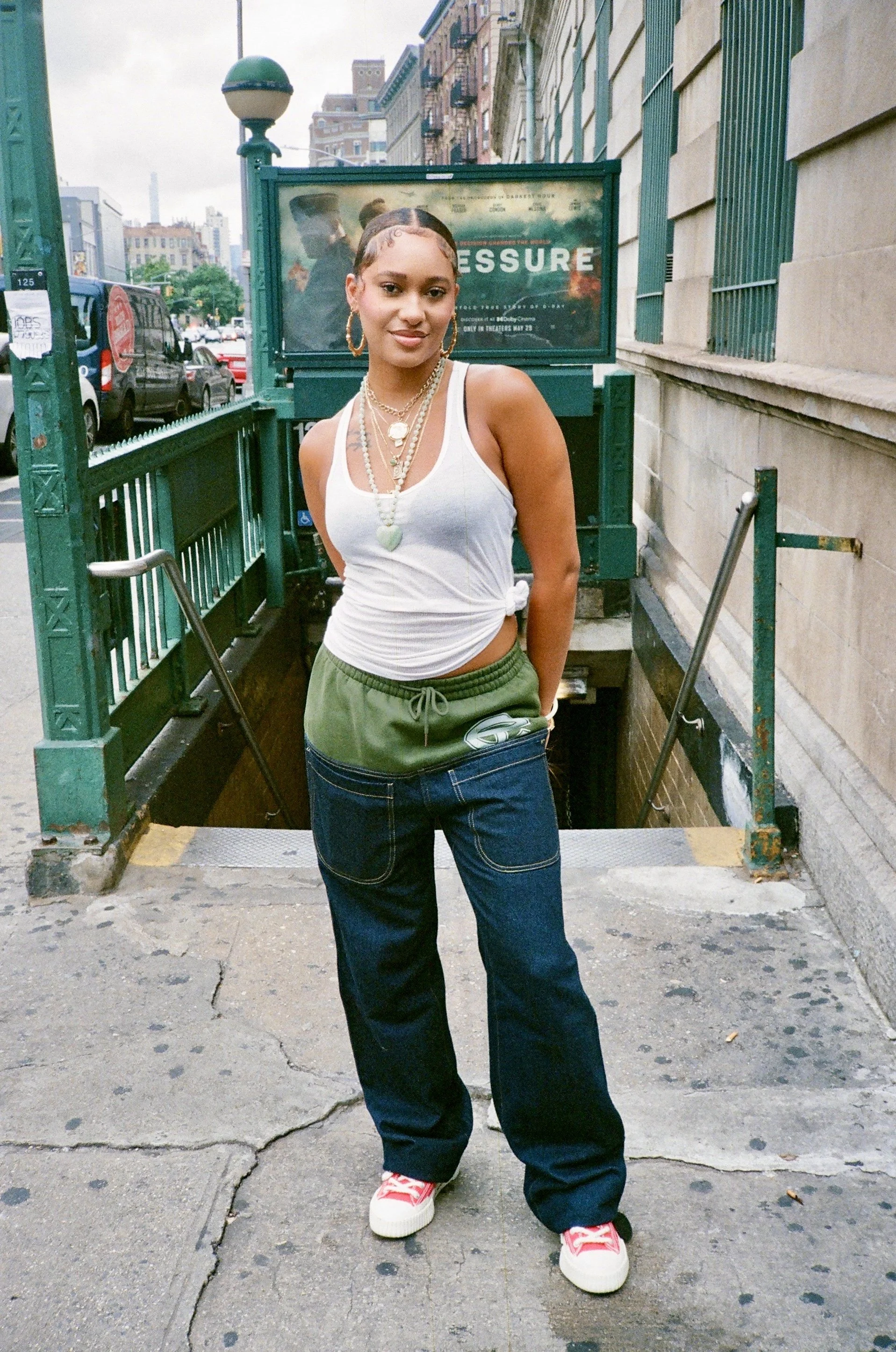 Young woman standing outside on a city street, wearing a white tank top, green sweatpants, red and white sneakers, layered gold necklaces, hoop earrings, and styled hair with baby hairs.